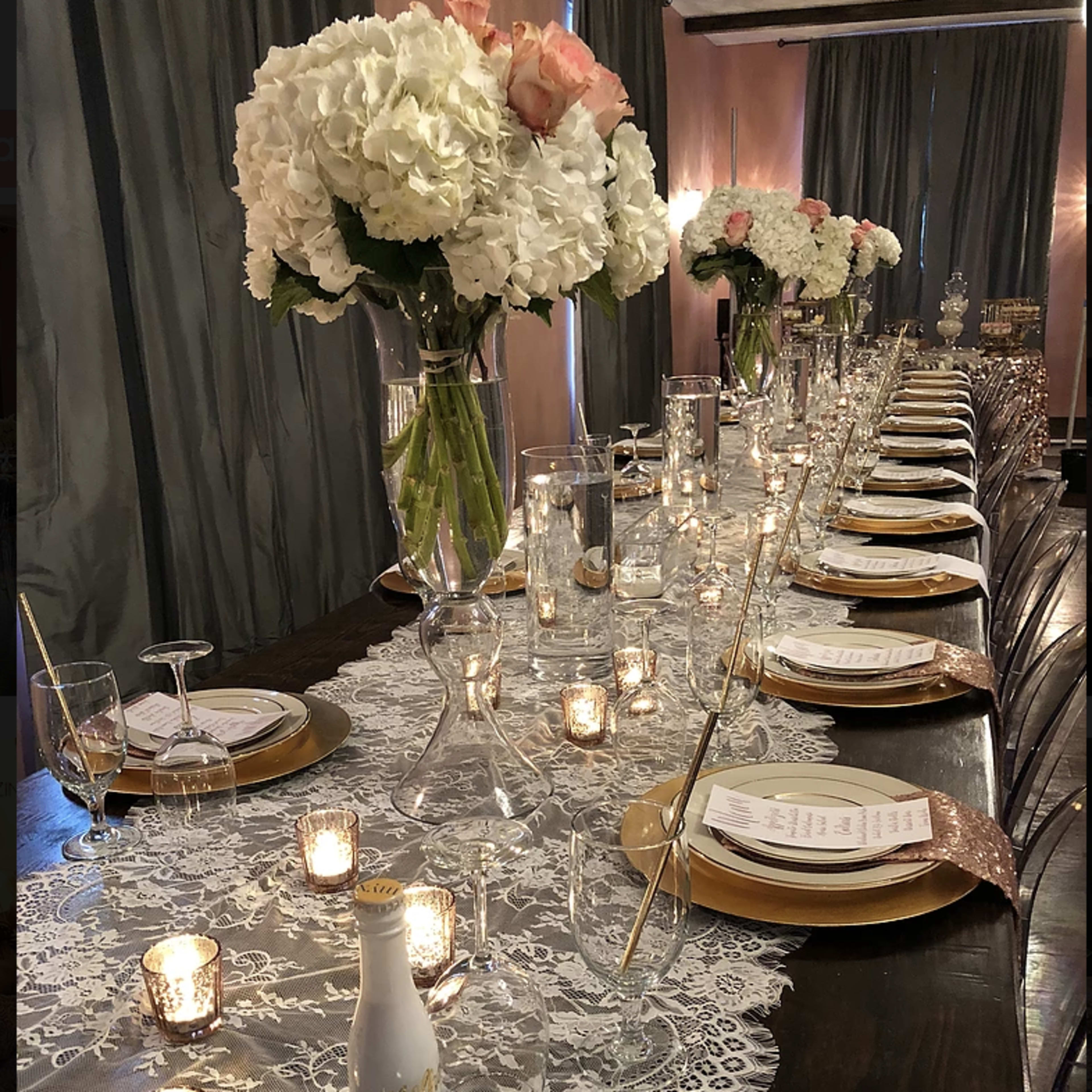 A long dining table is elegantly set with gold plates, white floral centerpieces, and lit candles alongside printed menus.