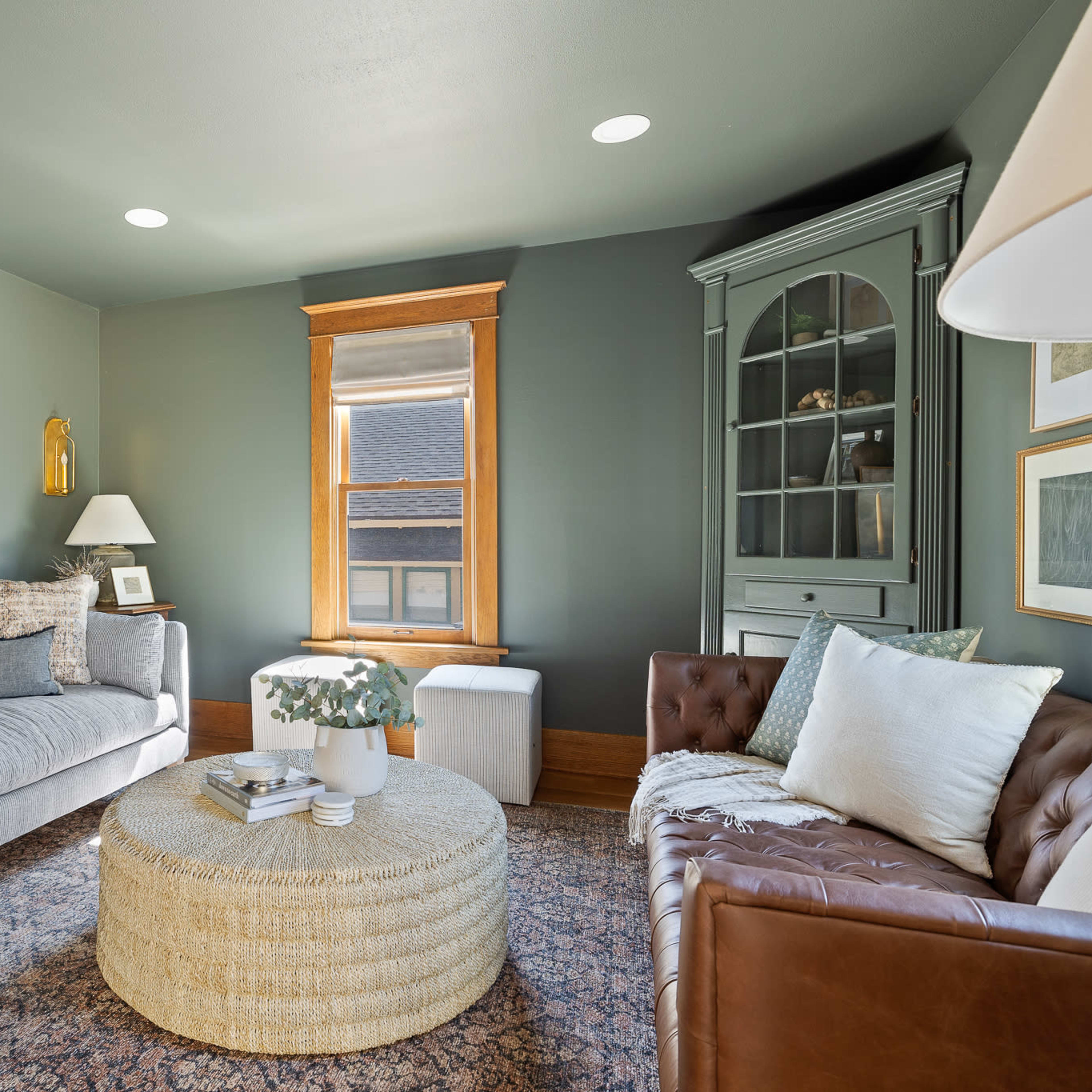 A cozy living room with a gray sofa, a brown leather couch, a round woven coffee table, and a window with natural light.