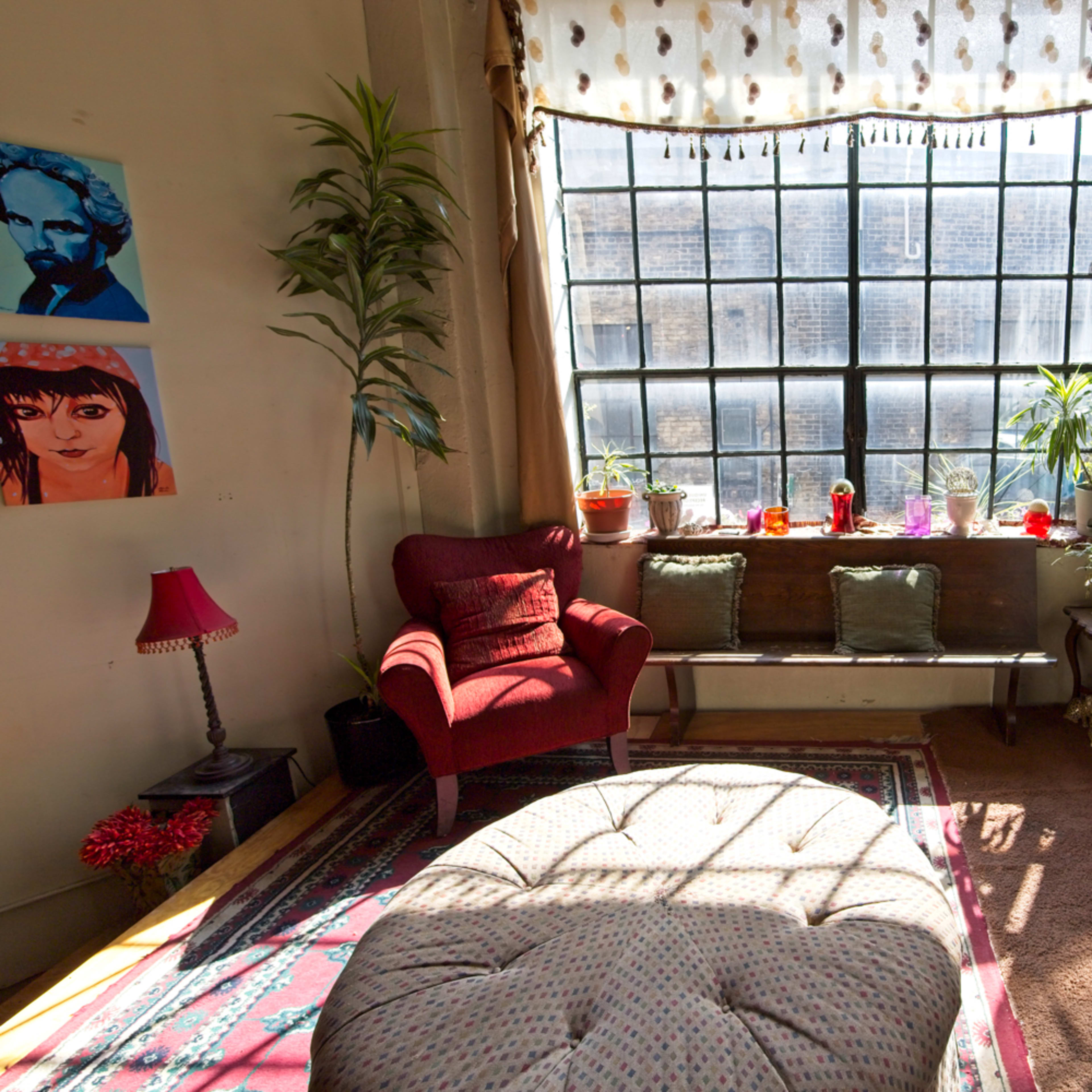 A cozy living room features a large window with plants and colorful decor, including a red chair and a round ottoman.