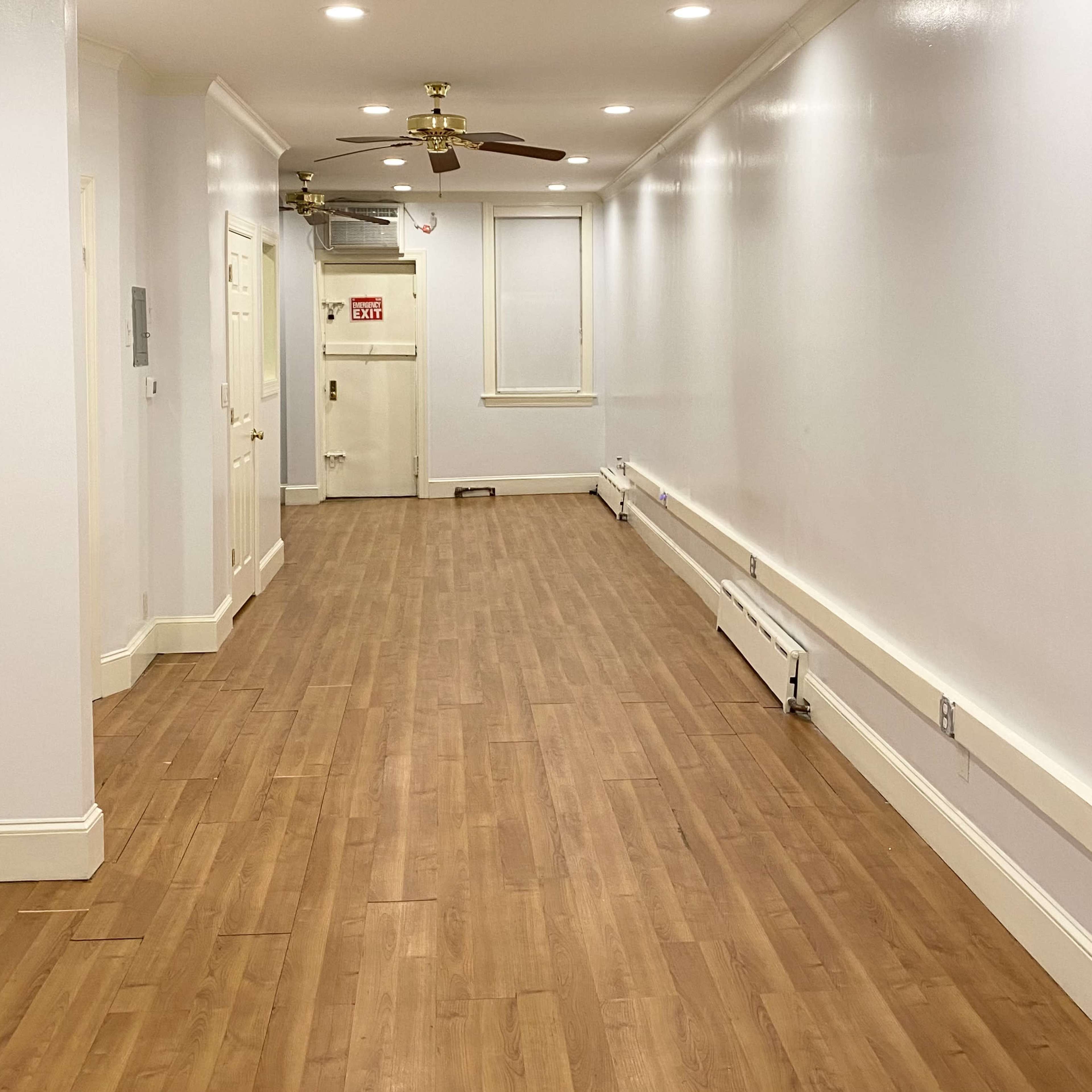 A long, narrow hallway with wooden flooring and white walls, featuring ceiling lights and a ceiling fan.
