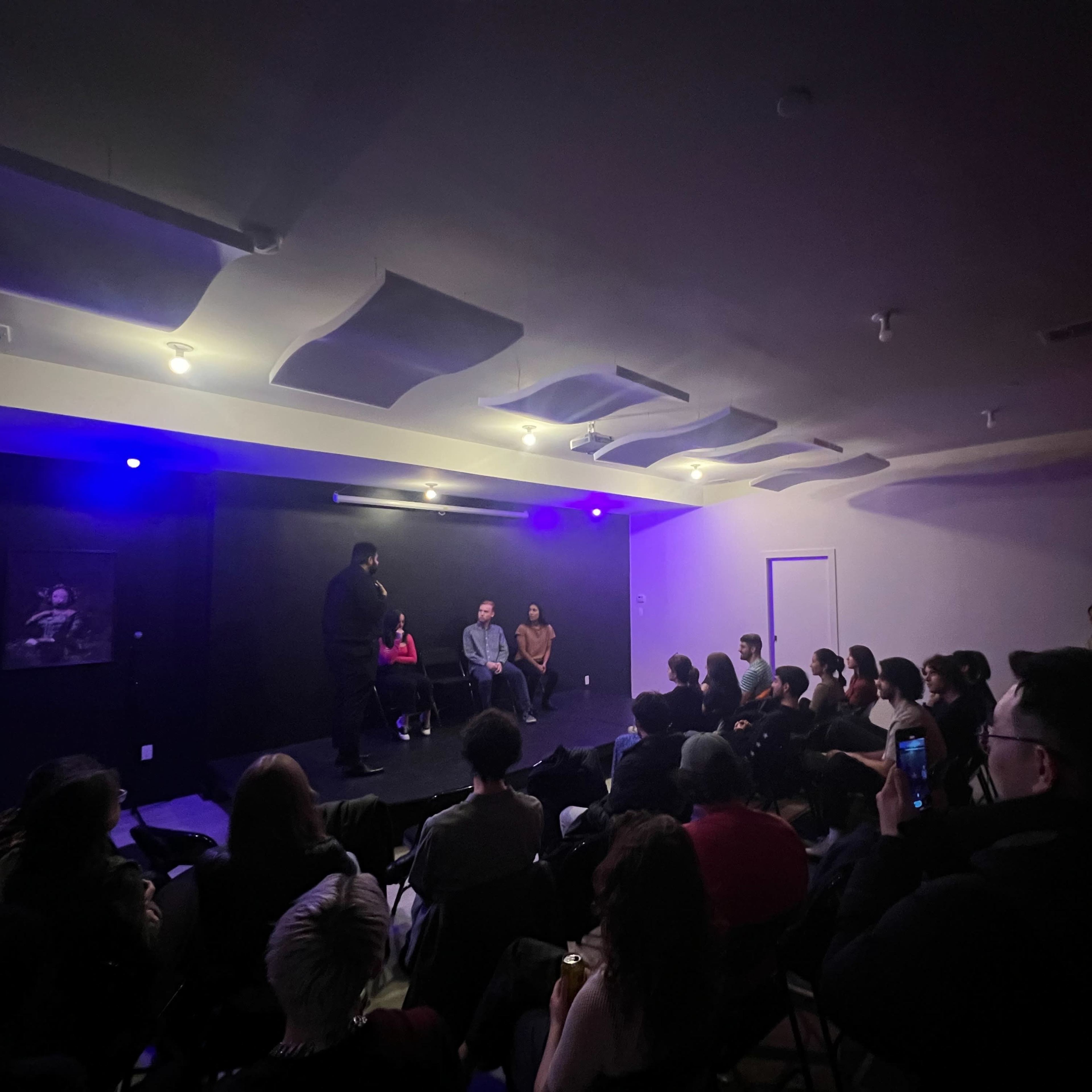 A seated audience watches a panel discussion on a small stage illuminated by dim blue lights.