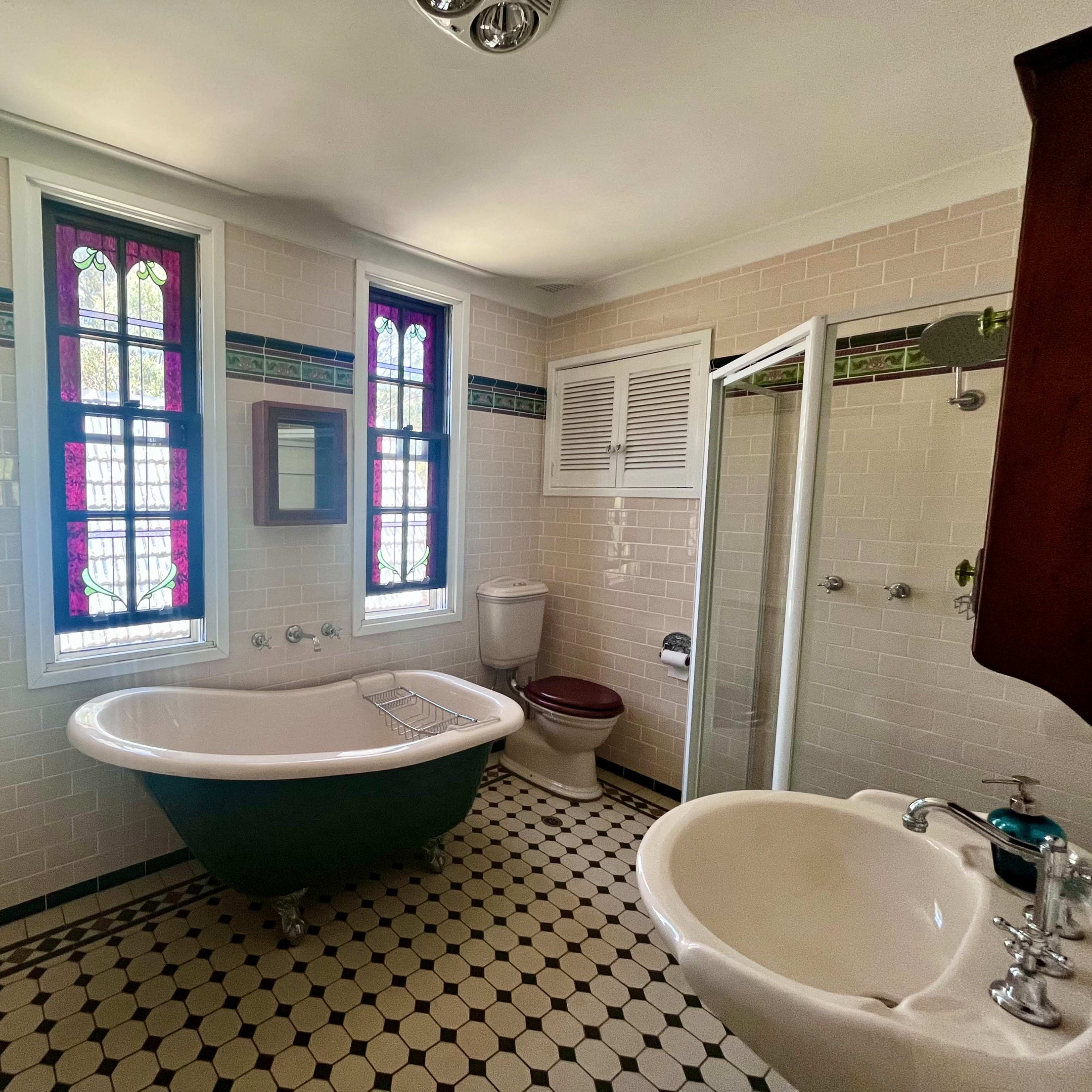 The bathroom features a freestanding clawfoot tub, a toilet, a sink, and two windows with stained glass, all set against a tiled floor and wall.