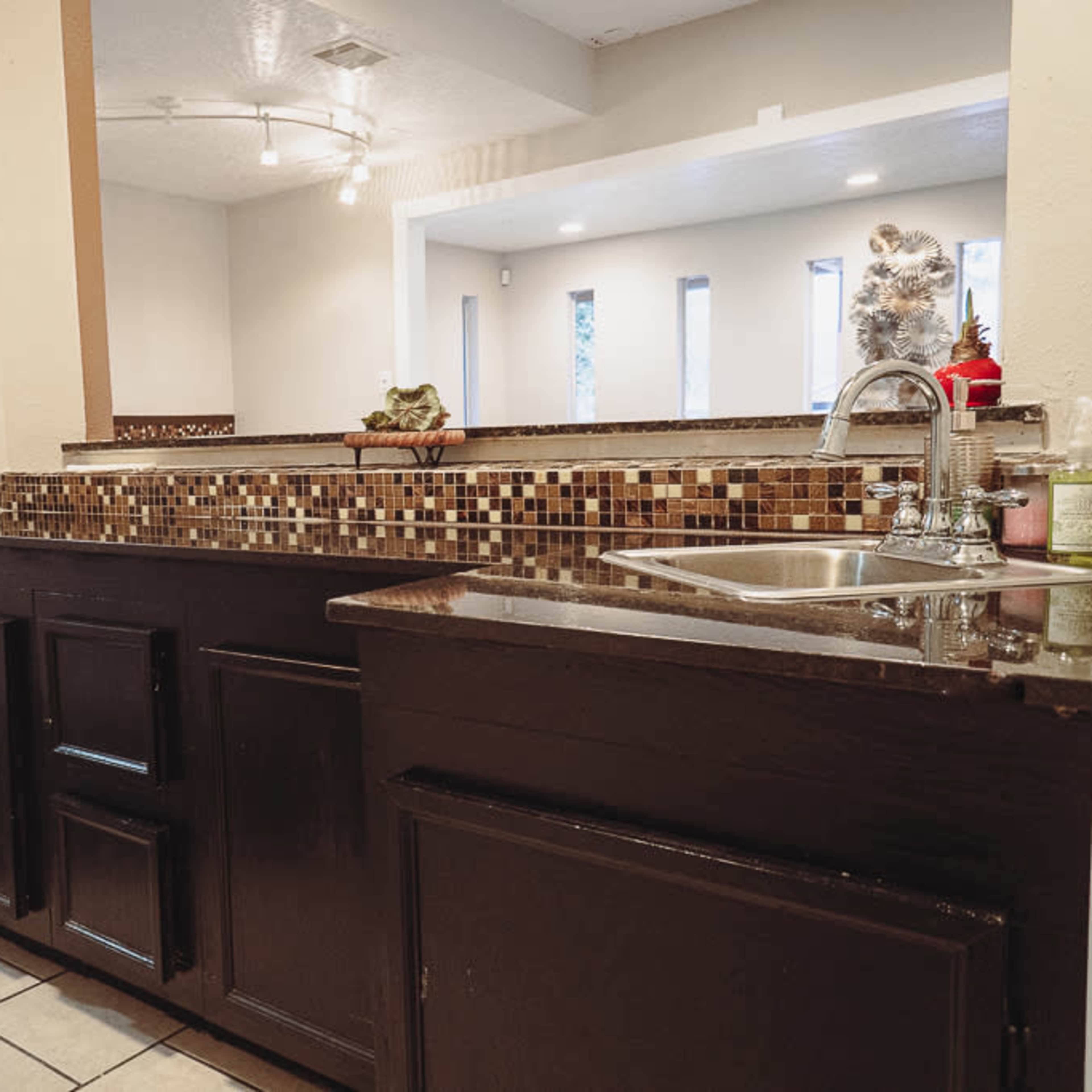 A kitchen area with dark wooden cabinetry, a sink, and a mosaic tile backsplash, leading to a bright living space with large windows.