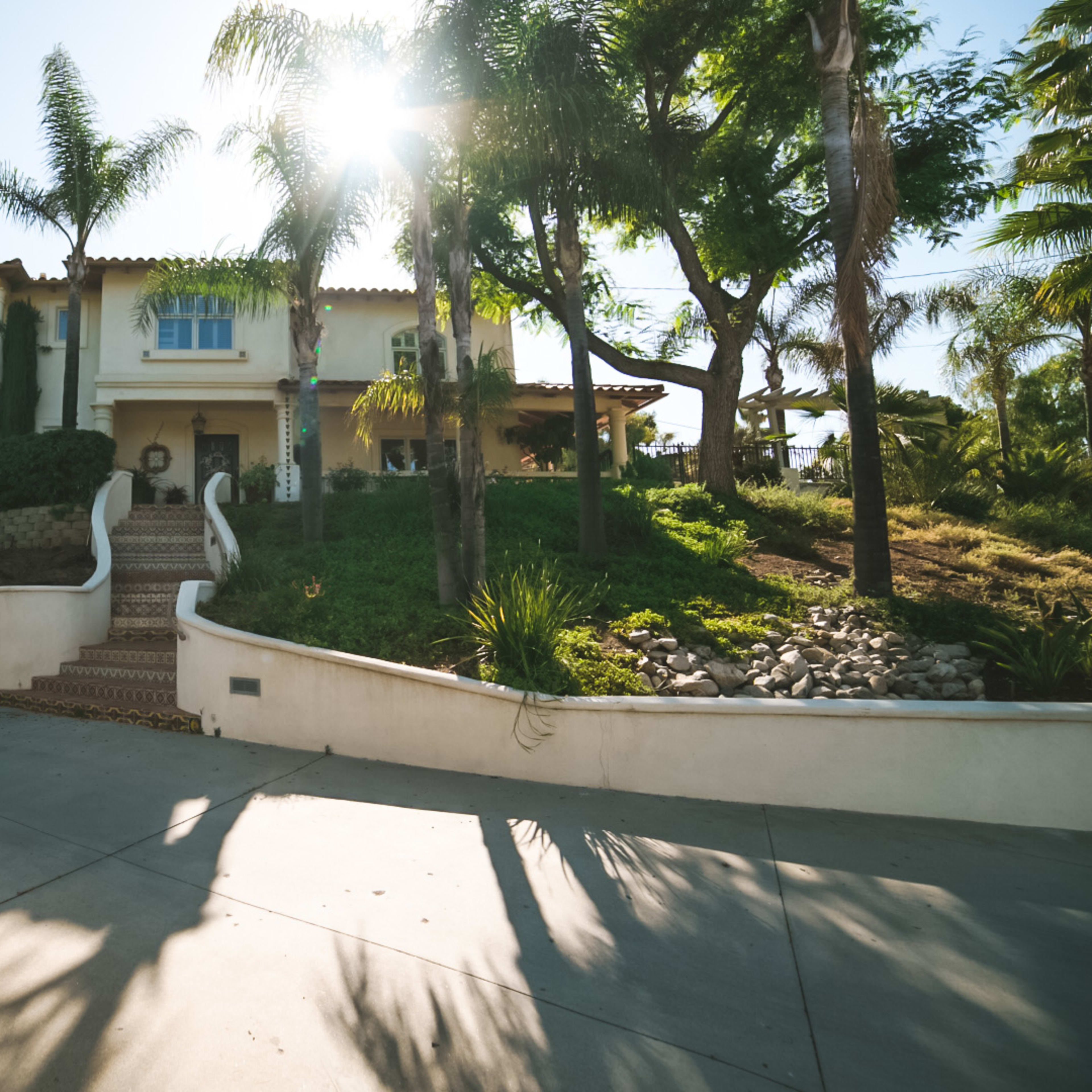 A large two-story house with palm trees and a landscaped yard is positioned on a sloped driveway under bright sunlight.
