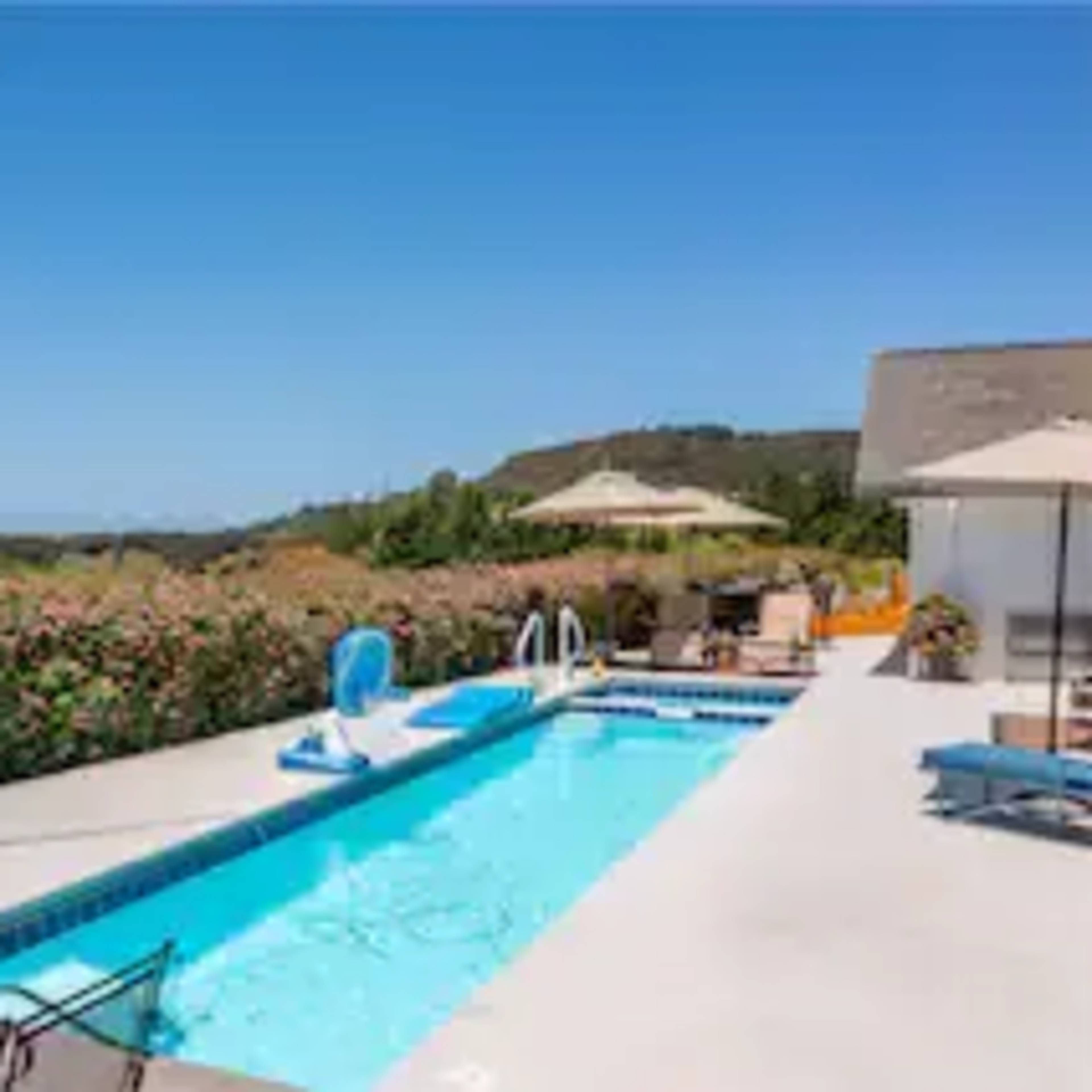A swimming pool is surrounded by lounge chairs and umbrellas, with a backdrop of lush greenery and hills.