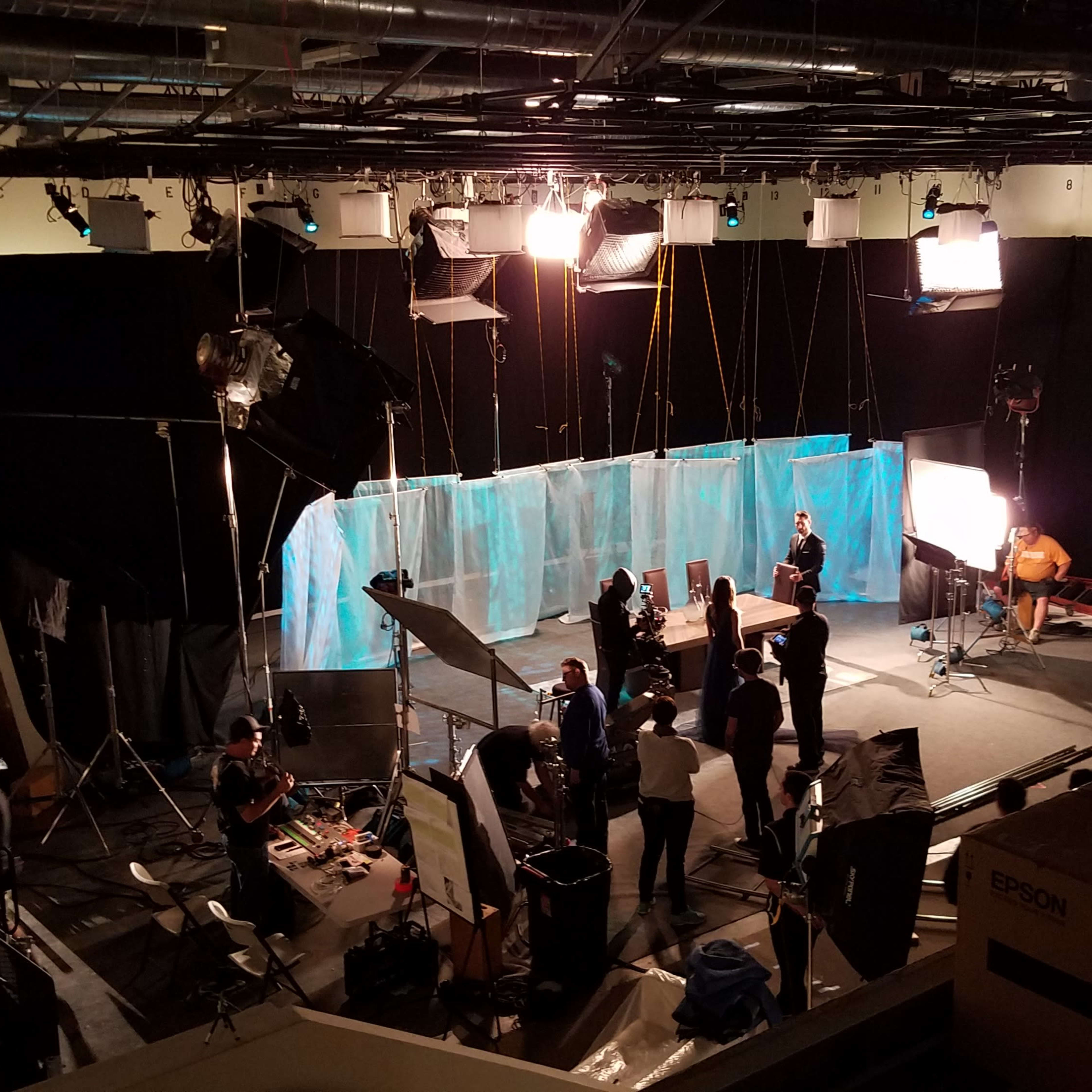 A film set is shown with crew members setting up equipment and lighting against a backdrop of blue and white screens.