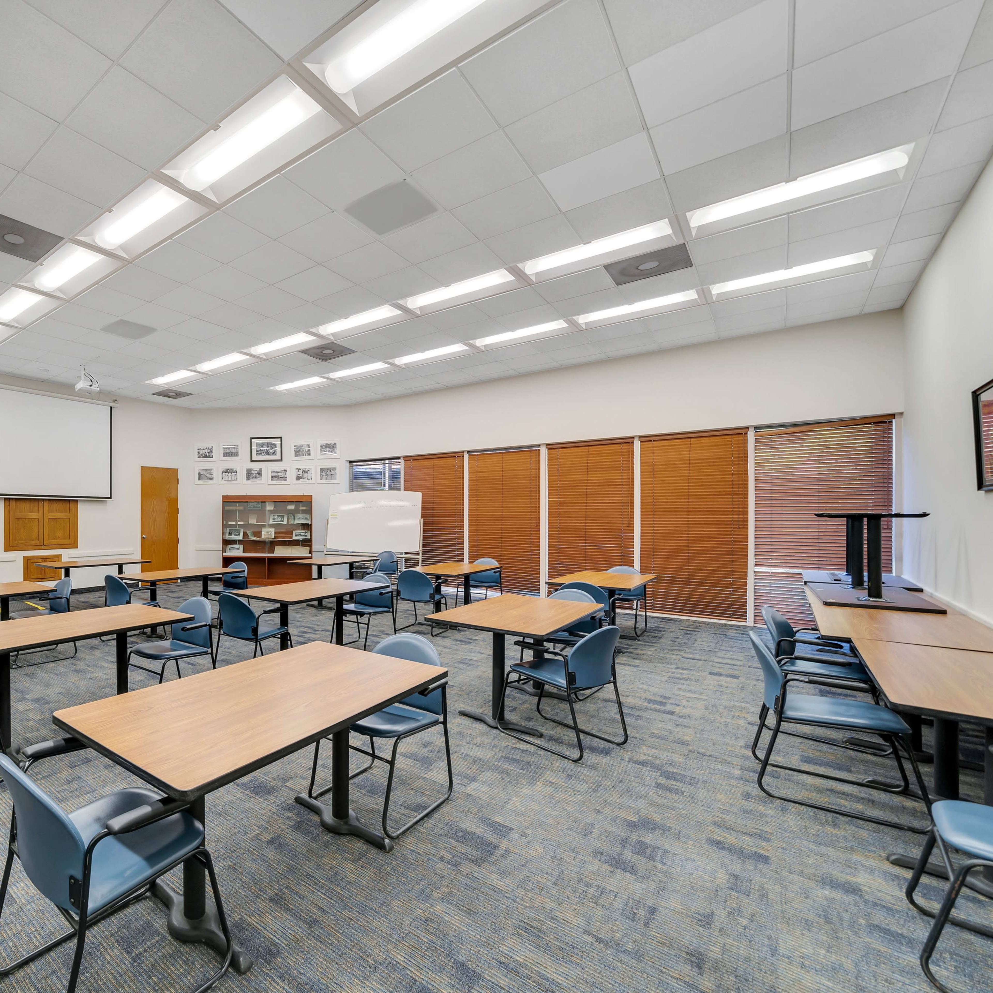 A classroom with several tables and chairs arranged, featuring a projector screen and multiple framed portraits on the walls.