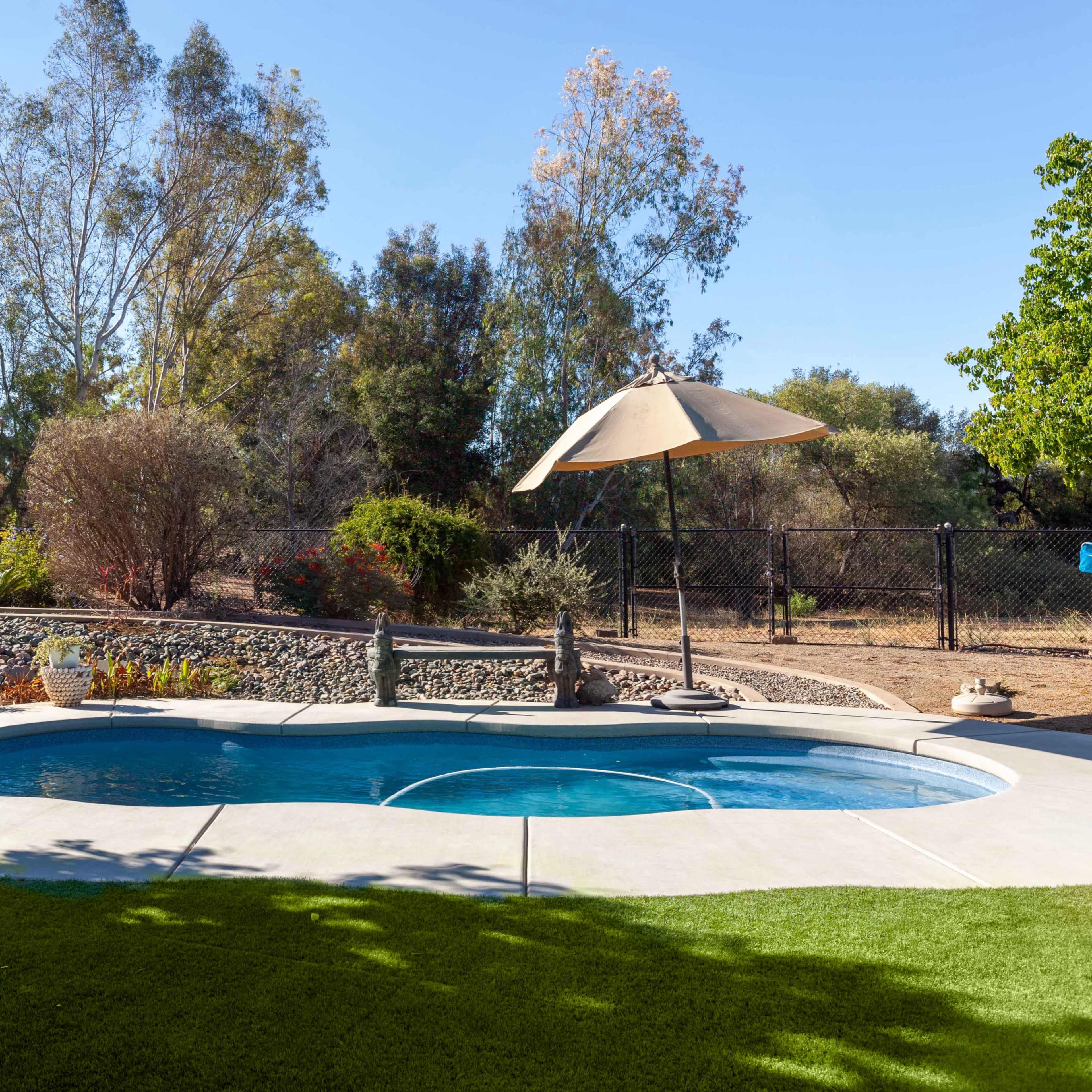 A backyard with a swimming pool, a patio umbrella, and landscaped greenery in the background.