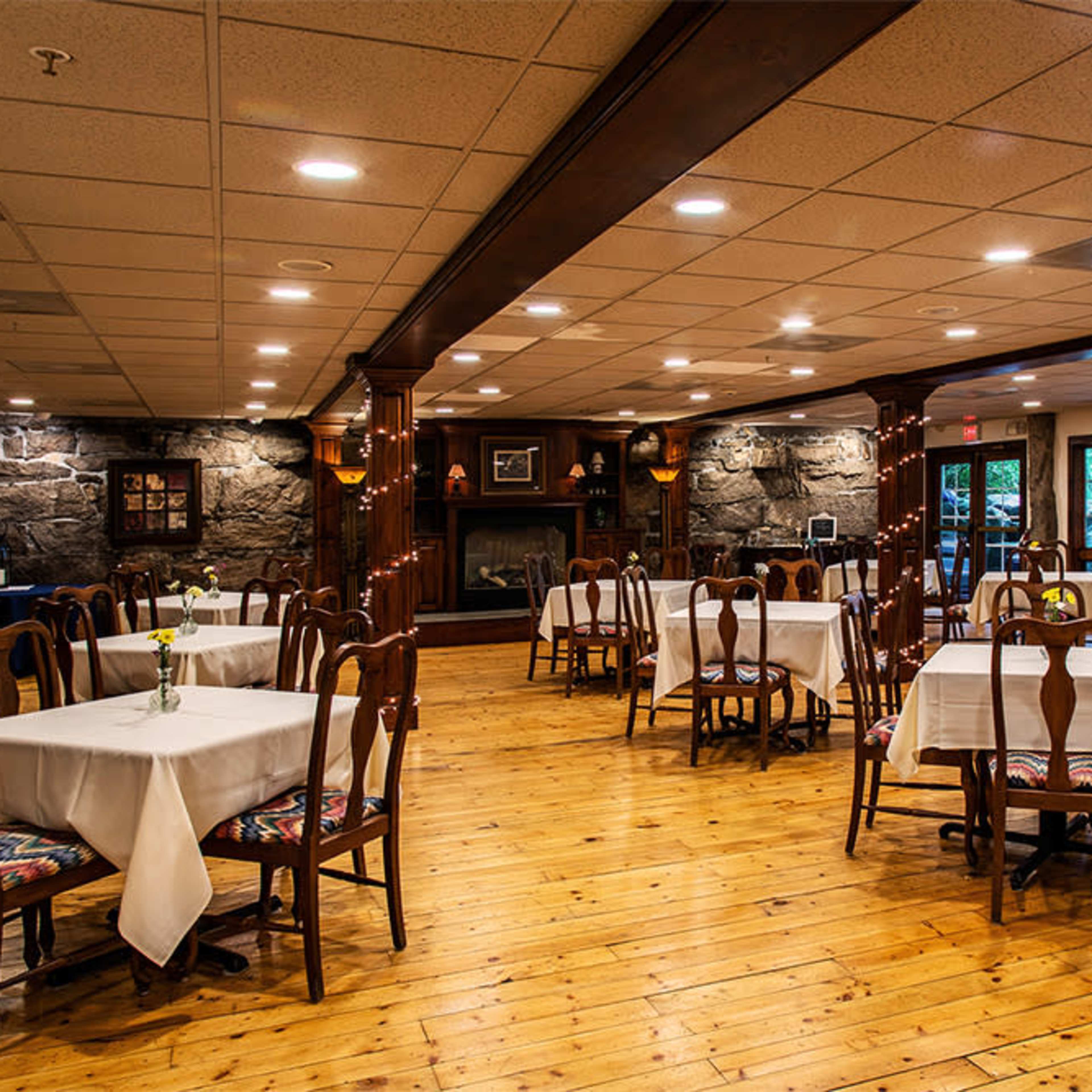 A spacious restaurant interior featuring wooden floors, several tables covered with white tablecloths, and a stone fireplace in the background.