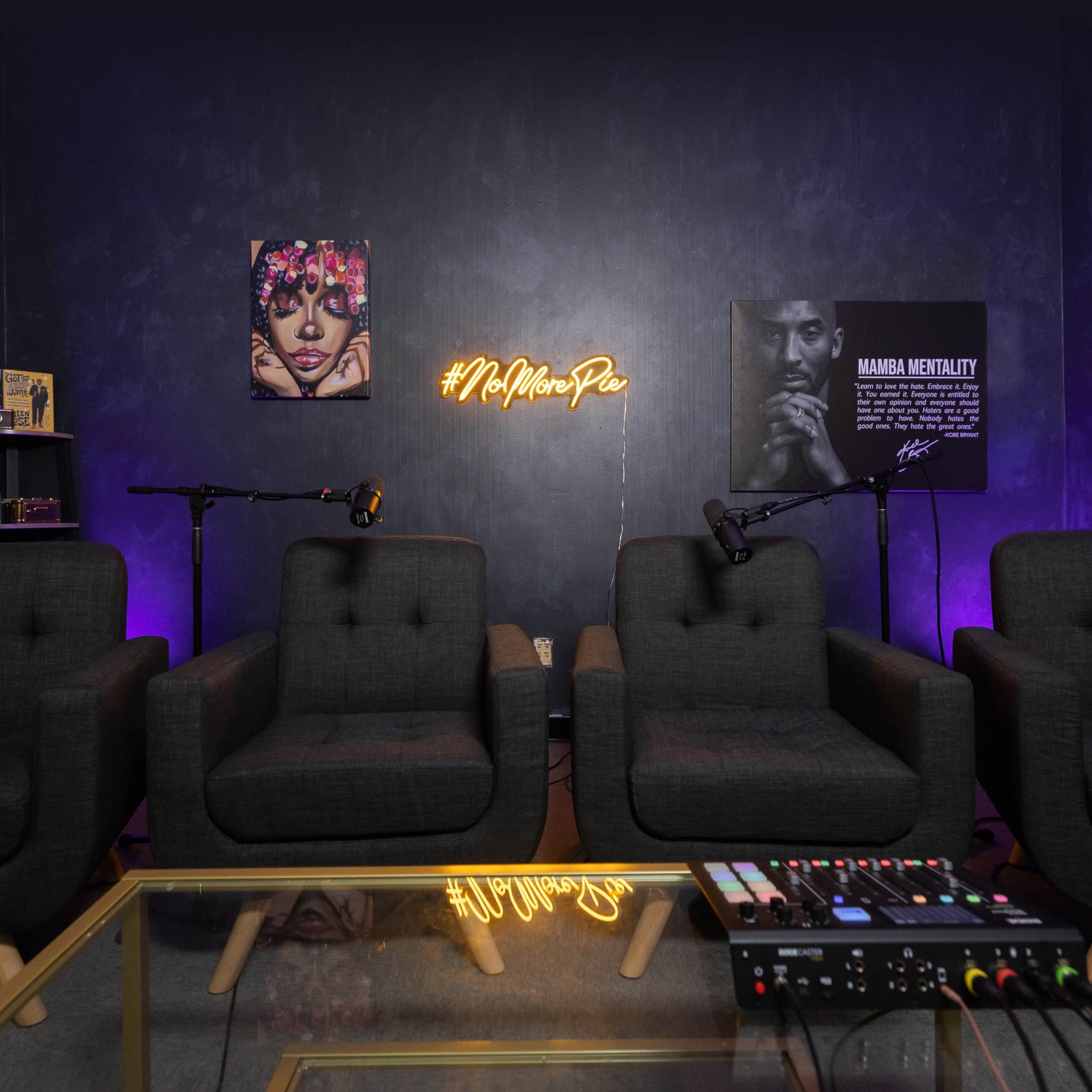 A small, modern podcast studio with four black chairs, a glass table, and two microphones, illuminated by a neon sign on the wall.
