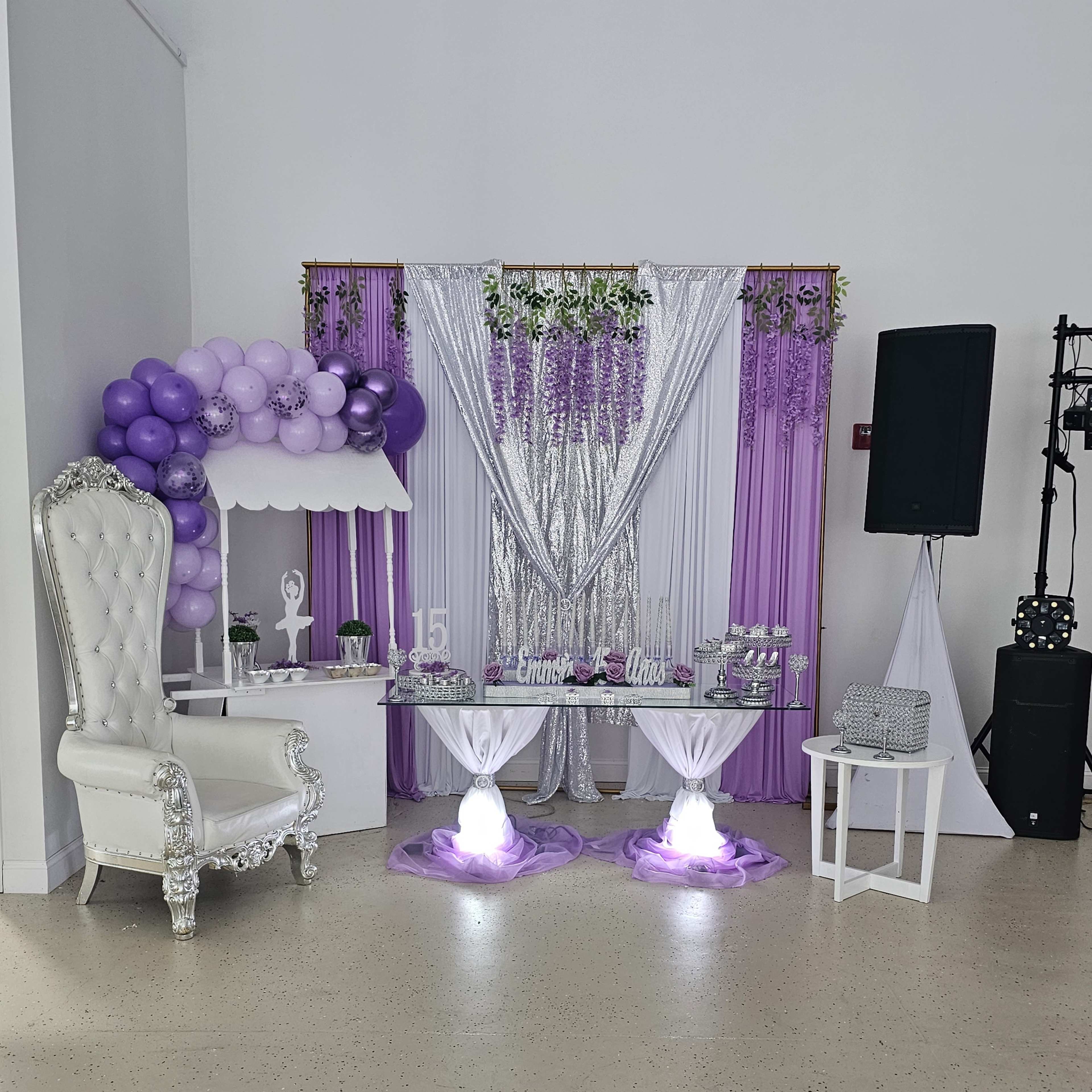 A decorated party area featuring a white throne chair, a purple and silver backdrop, a table setup with decorative items, and a balloon arch.