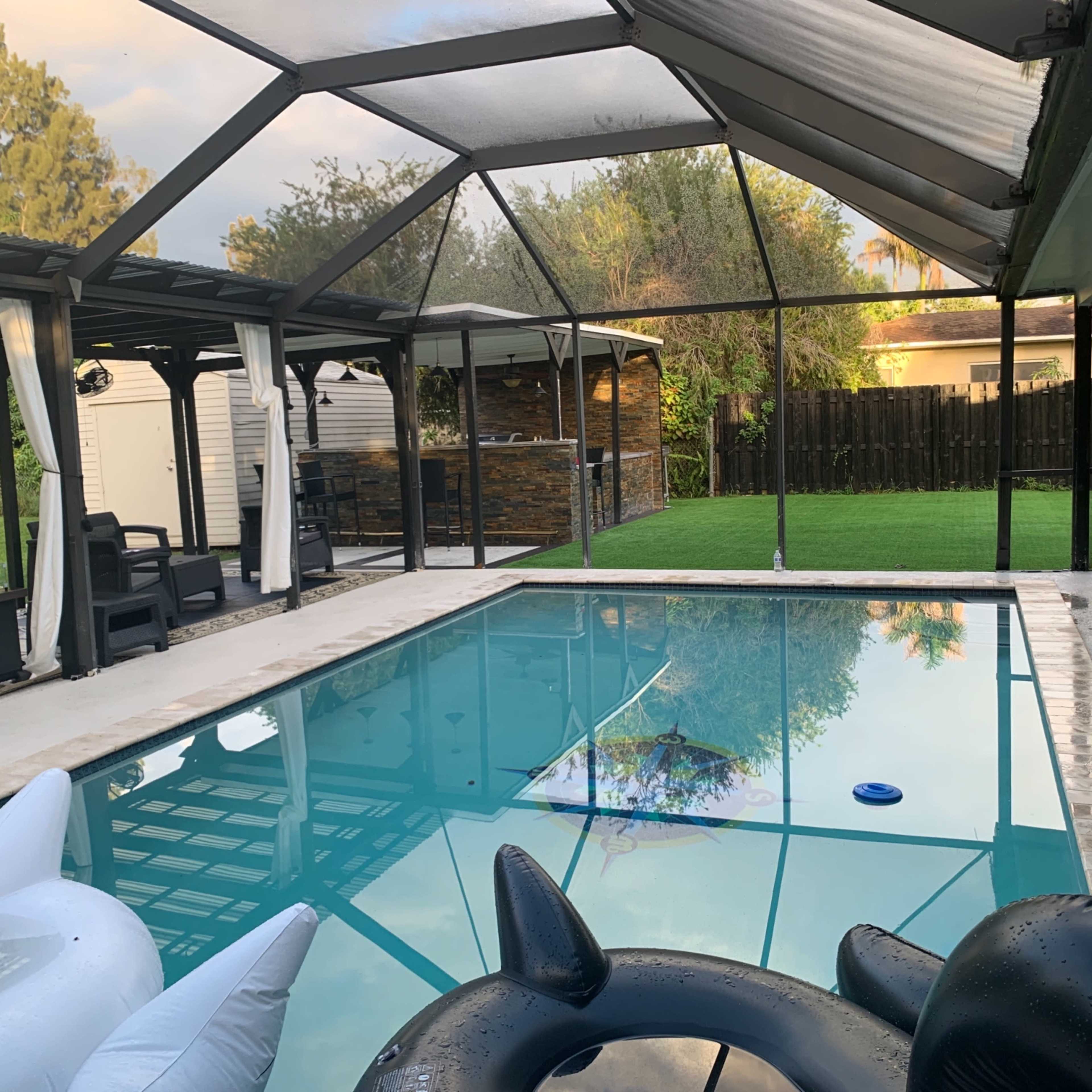 A swimming pool surrounded by a screened patio, with inflatable pool floats and a grassy area in the background.