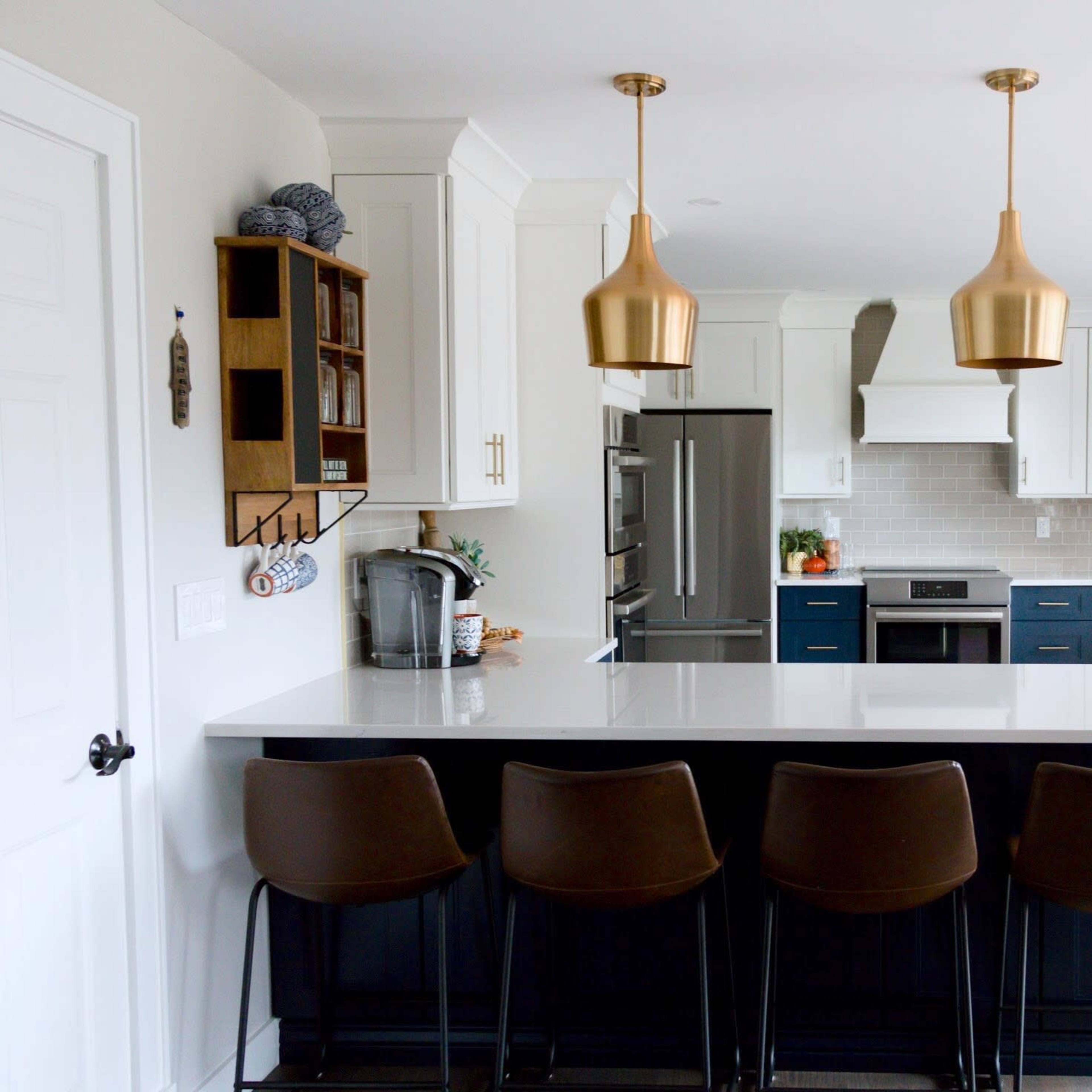 A modern kitchen features a large island with three brown bar stools, gold pendant lights, white upper cabinets, and navy lower cabinets.