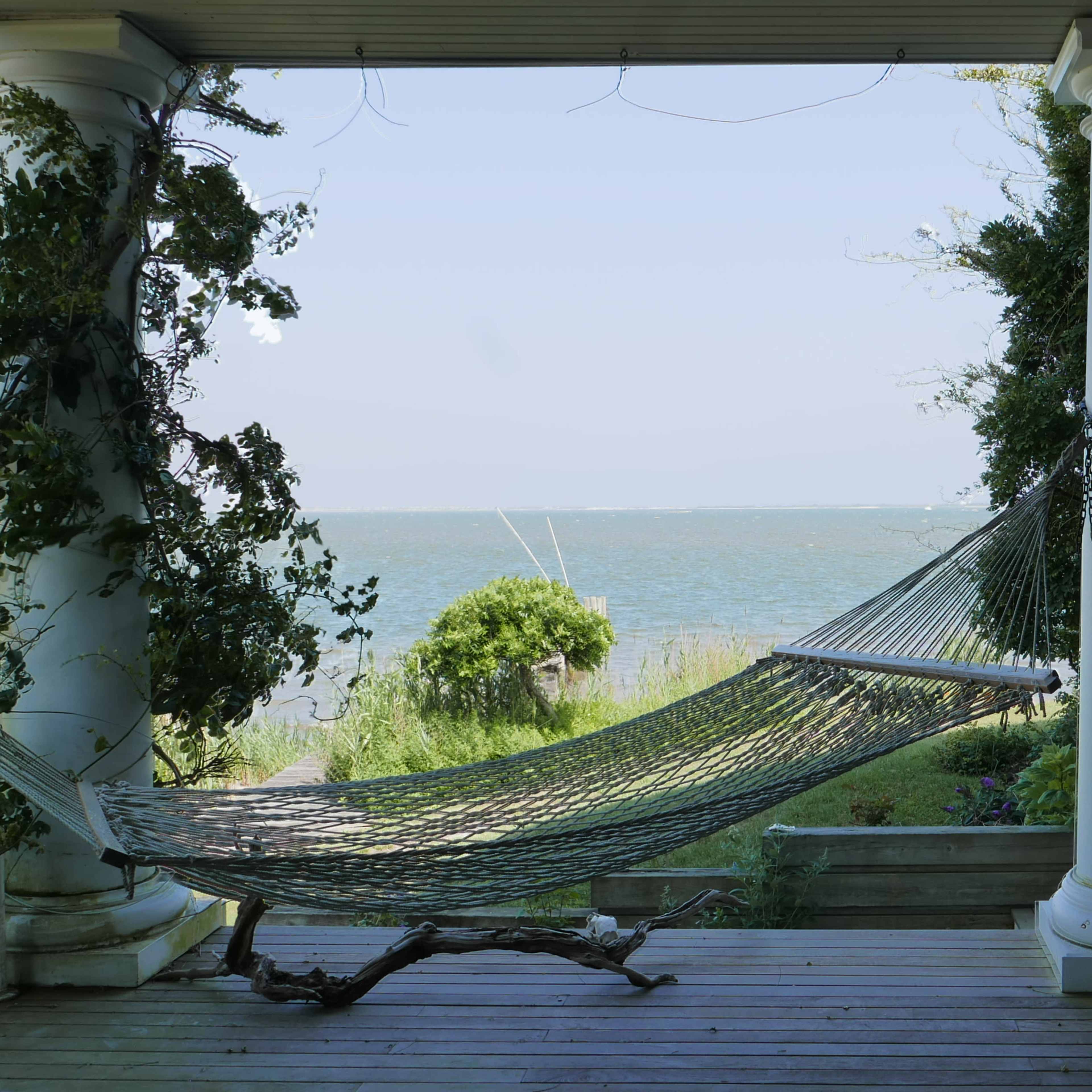 A hammock is suspended between two columns on a porch overlooking a calm body of water.