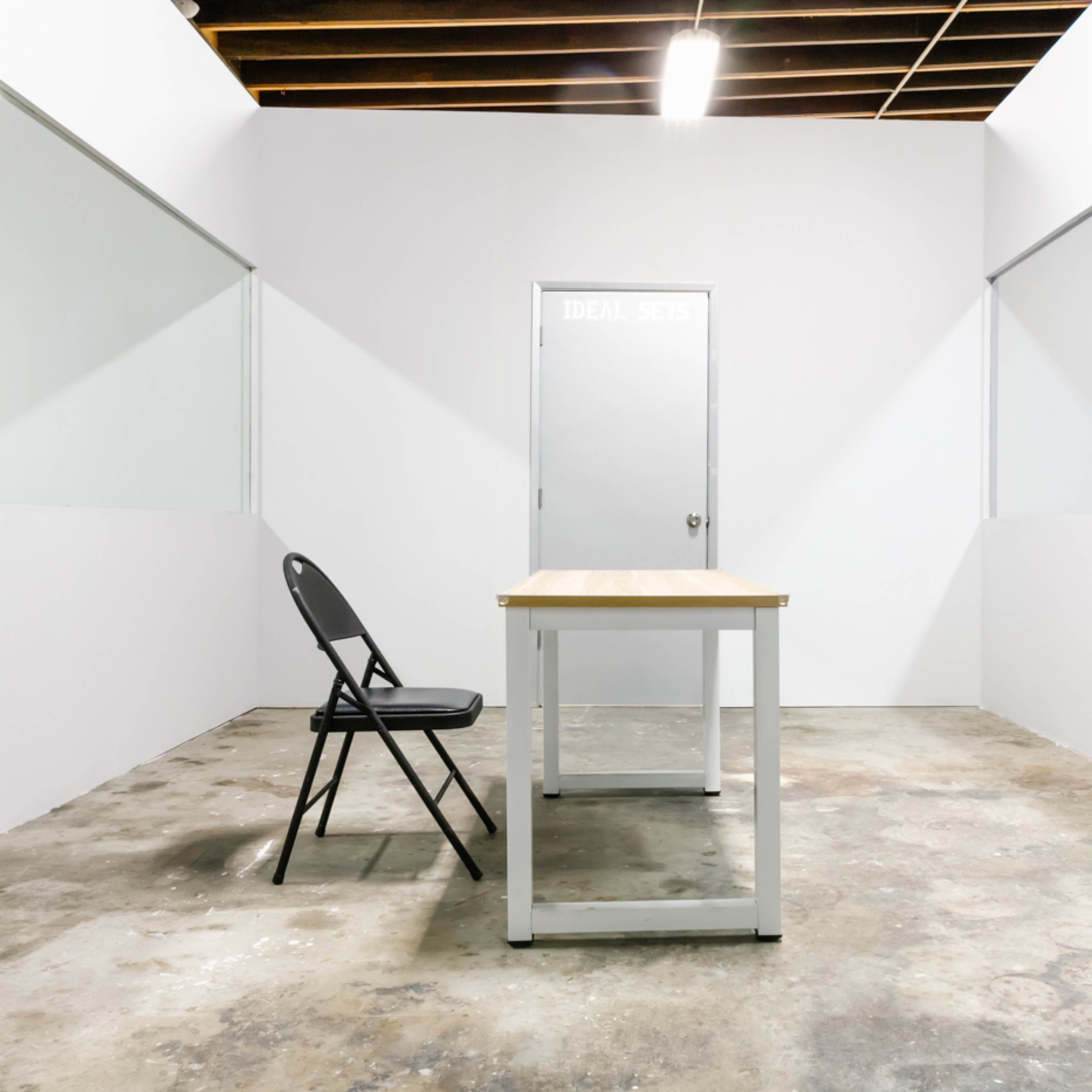 An empty room features a table and a chair in the center, with white walls and two glass panels on opposing sides.