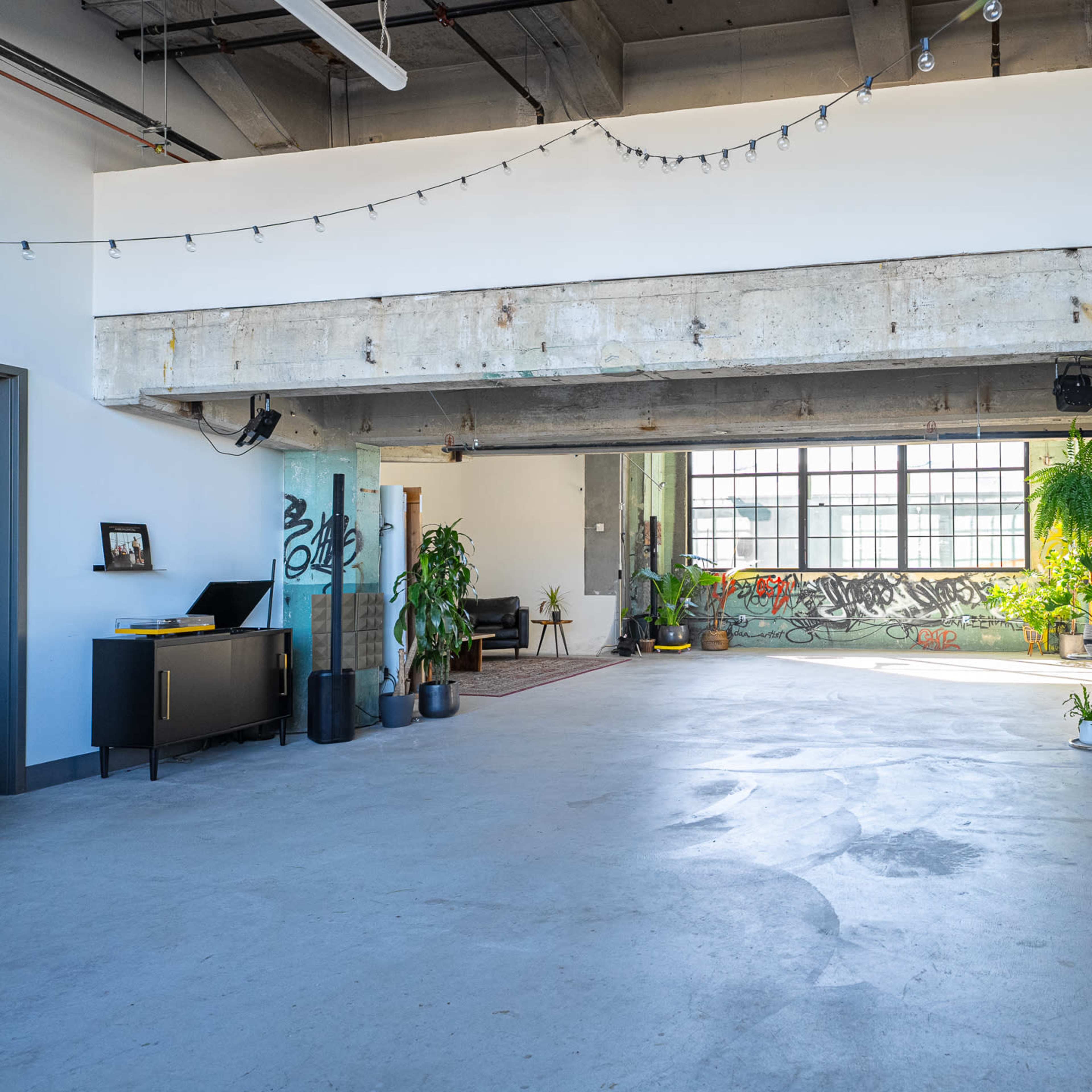 An open, industrial-style interior space with large windows, polished concrete floors, and greenery alongside a graffiti-covered wall.