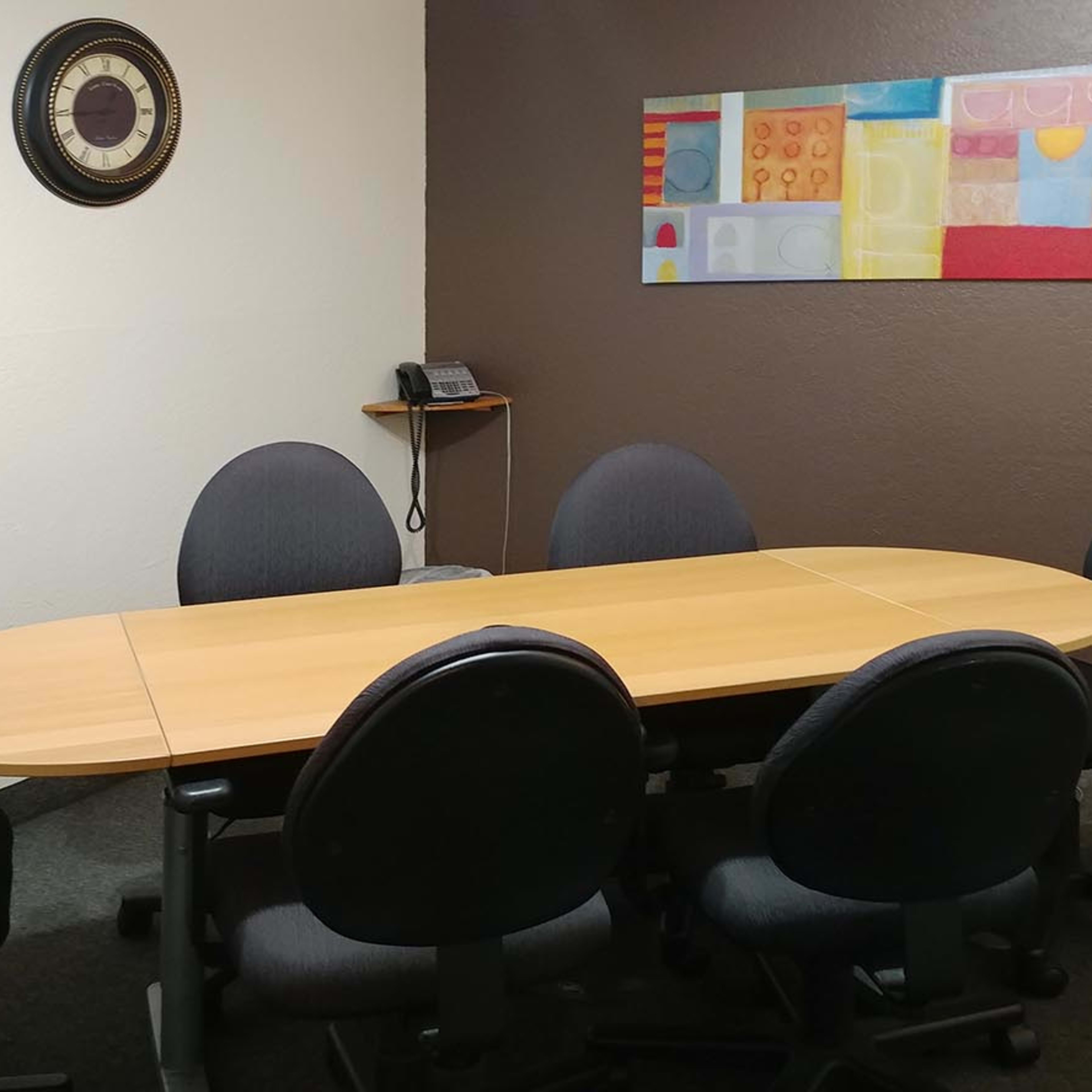 A conference room features an oval wooden table surrounded by six rolling chairs, with a wall-mounted phone and abstract artwork in the background.