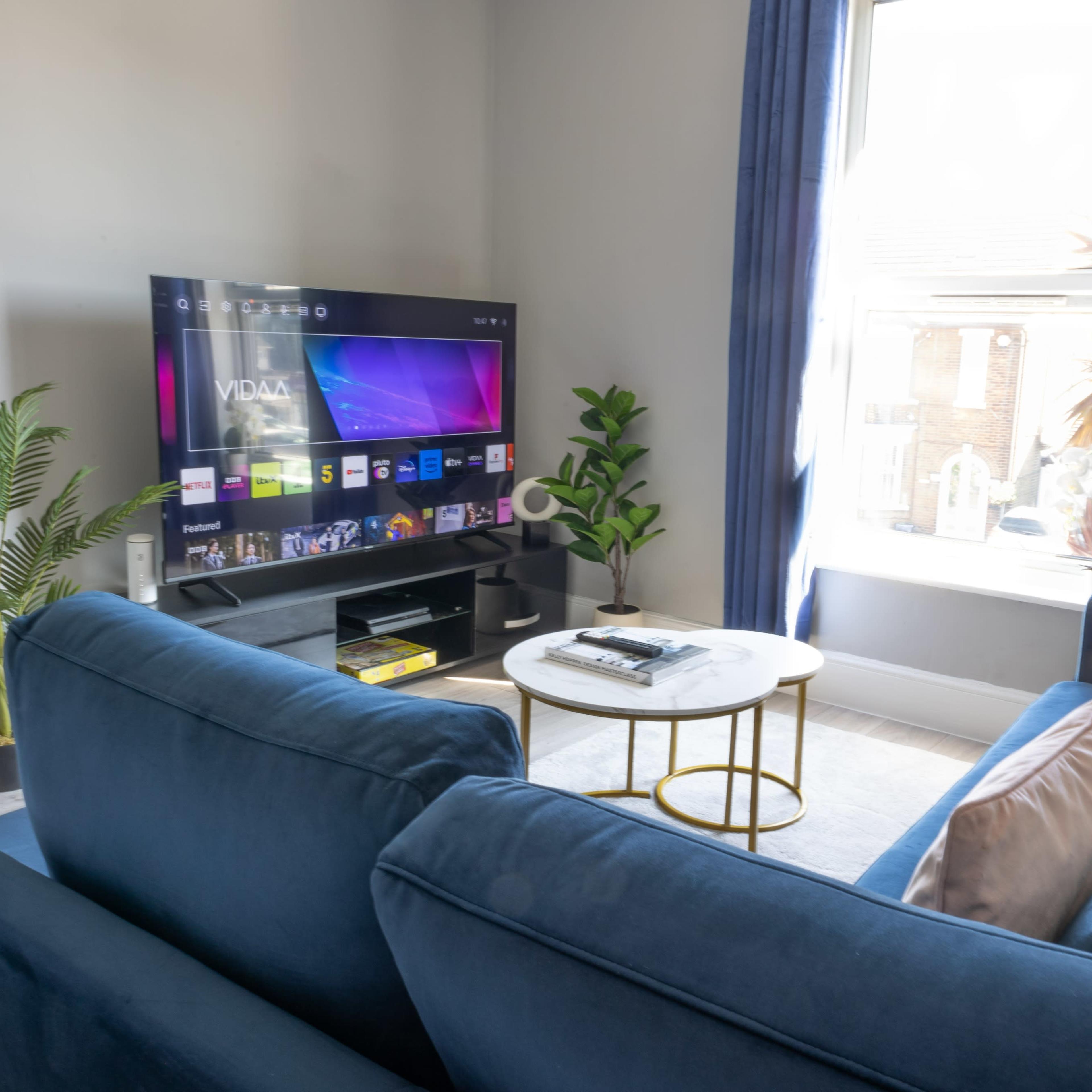 A living room with a blue sofa, a round coffee table, a television displaying a menu, and a potted plant near a window.