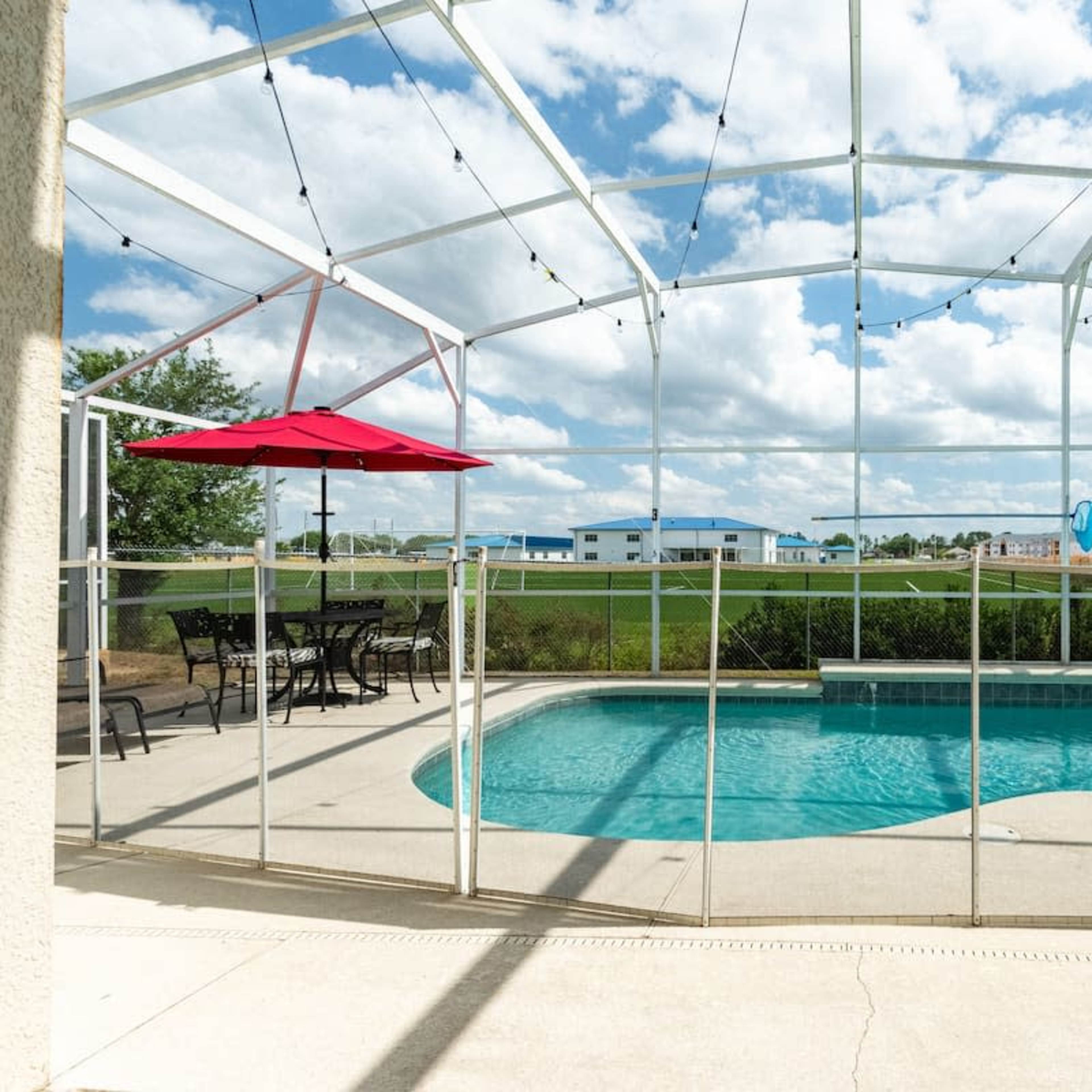 A screened-in pool area with a clear blue pool, a red umbrella over a dining set, and a grassy field visible in the background under a partly cloudy sky.