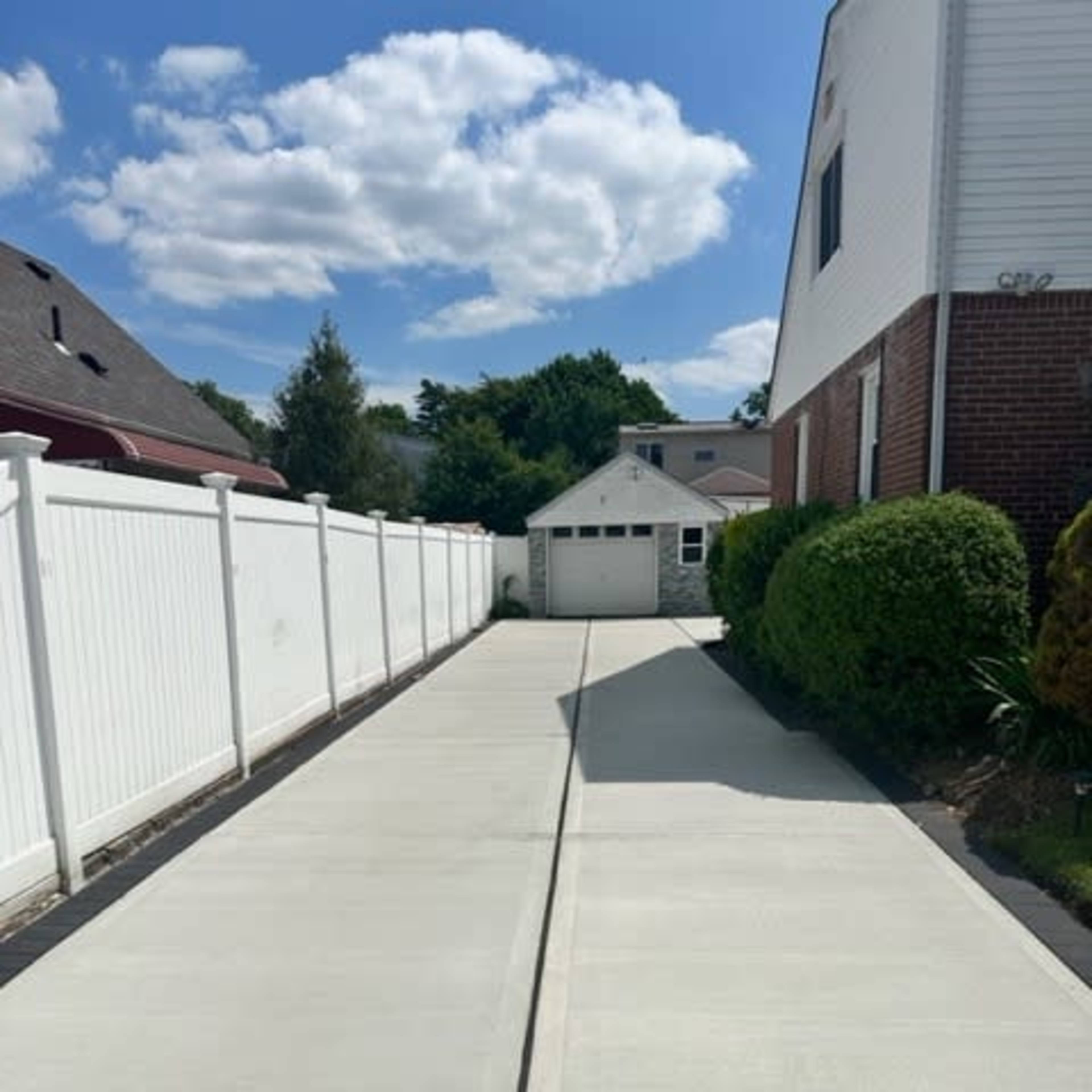 A concrete driveway leads to a garage at the end, bordered by a white fence and greenery on both sides.
