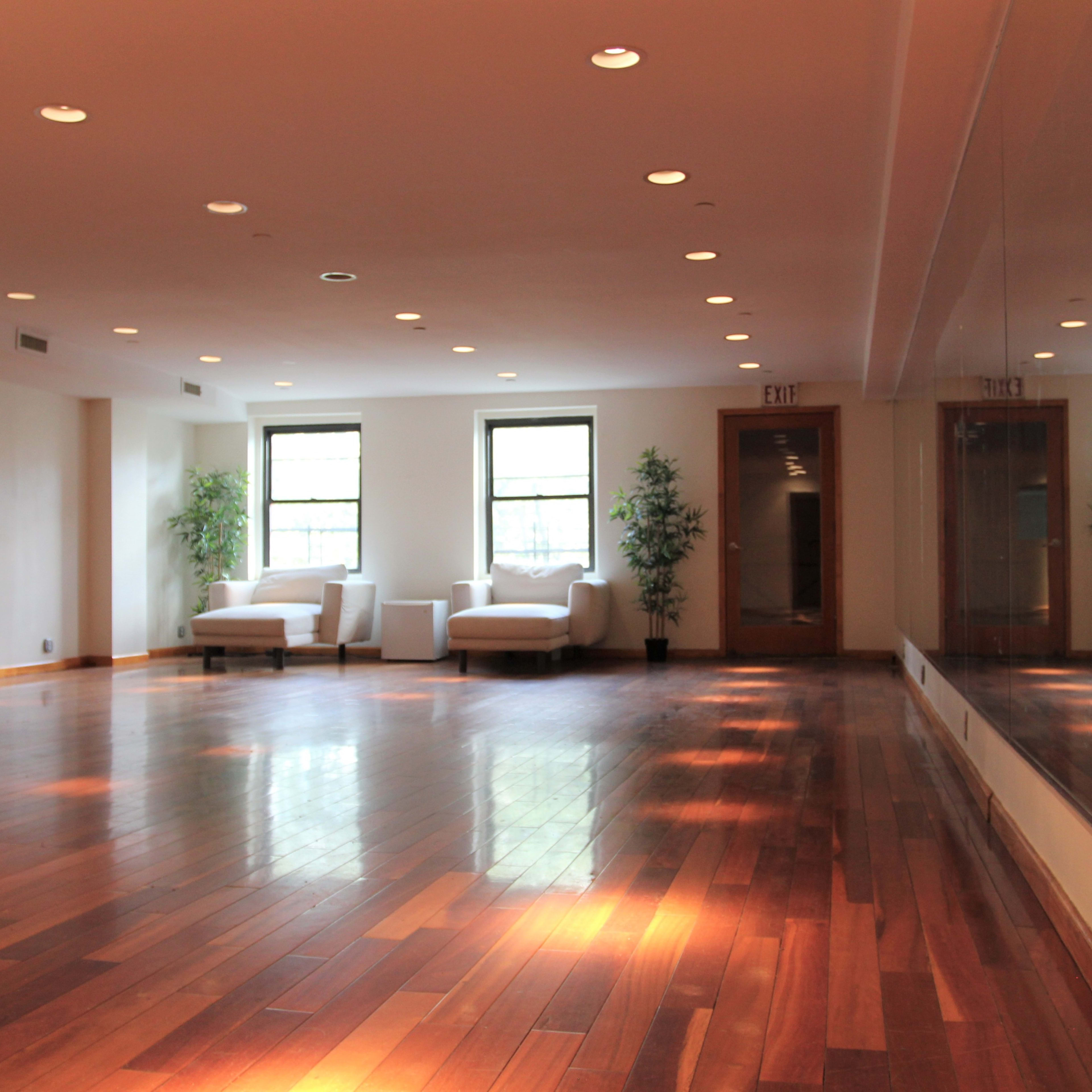 A spacious room with polished wooden floors, large windows, and two white couches against the walls.