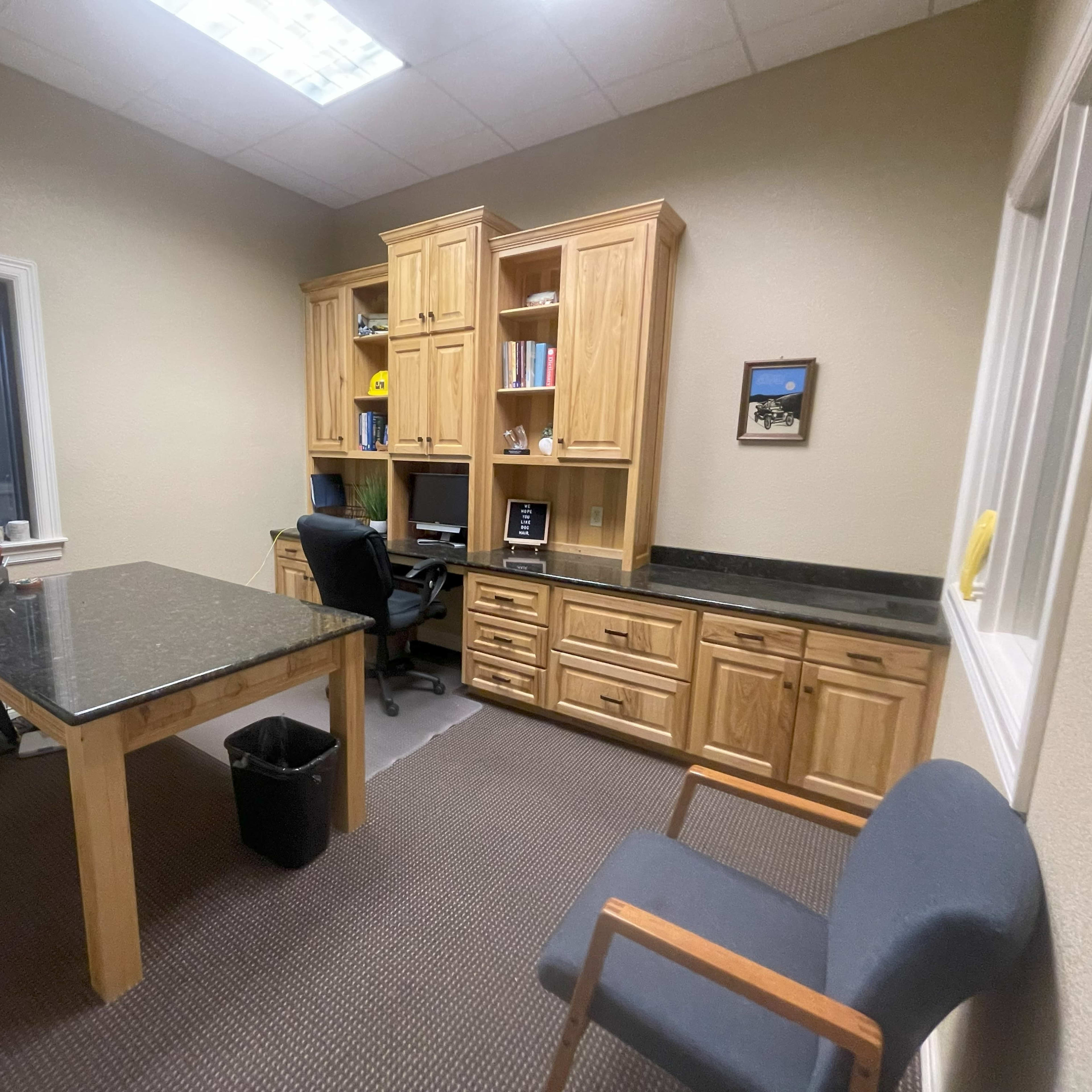 A spacious office featuring a wooden desk with a black top, a matching cabinet filled with books, and a single chair beside a window.