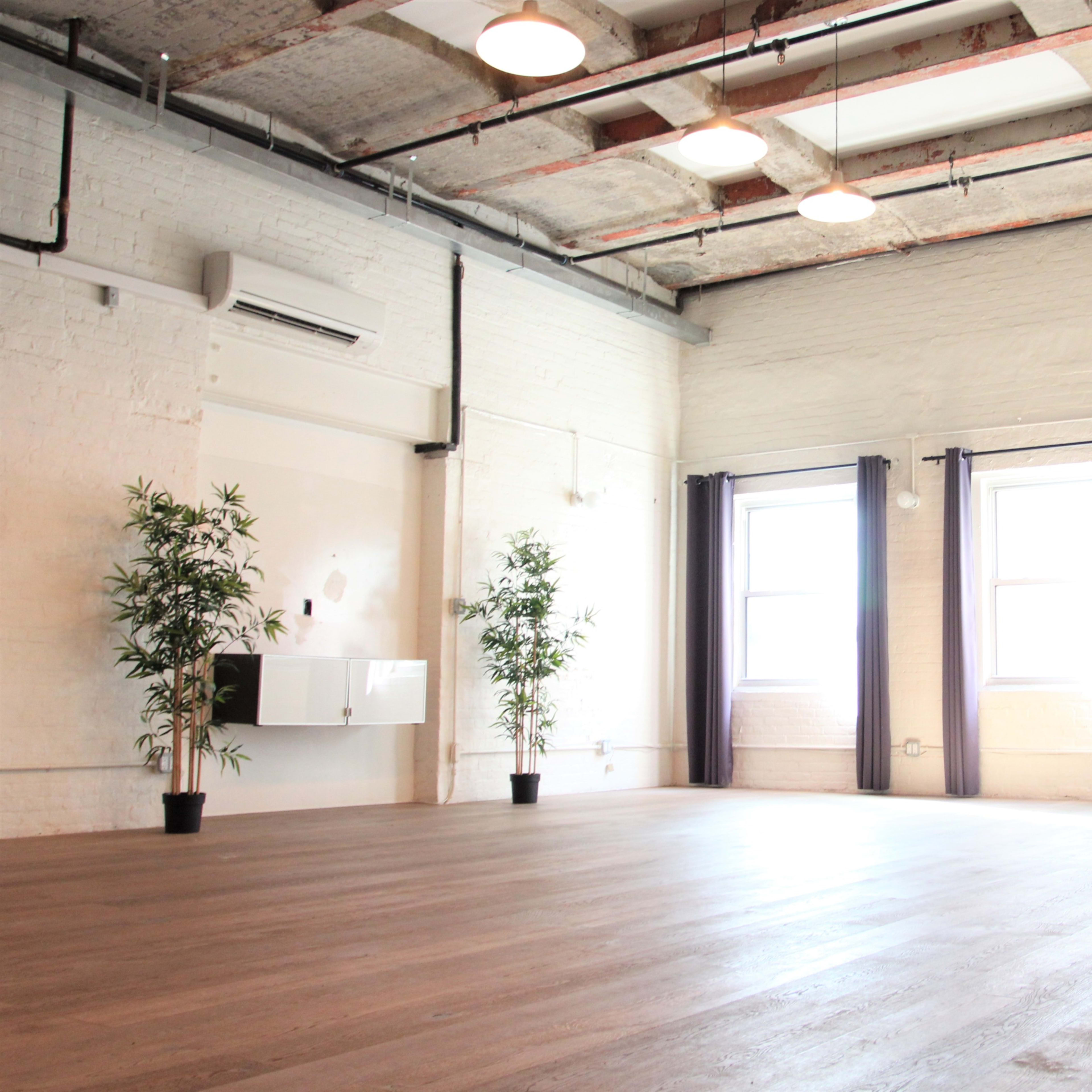 A spacious, empty room with wooden flooring, large windows, and potted plants in the corners.