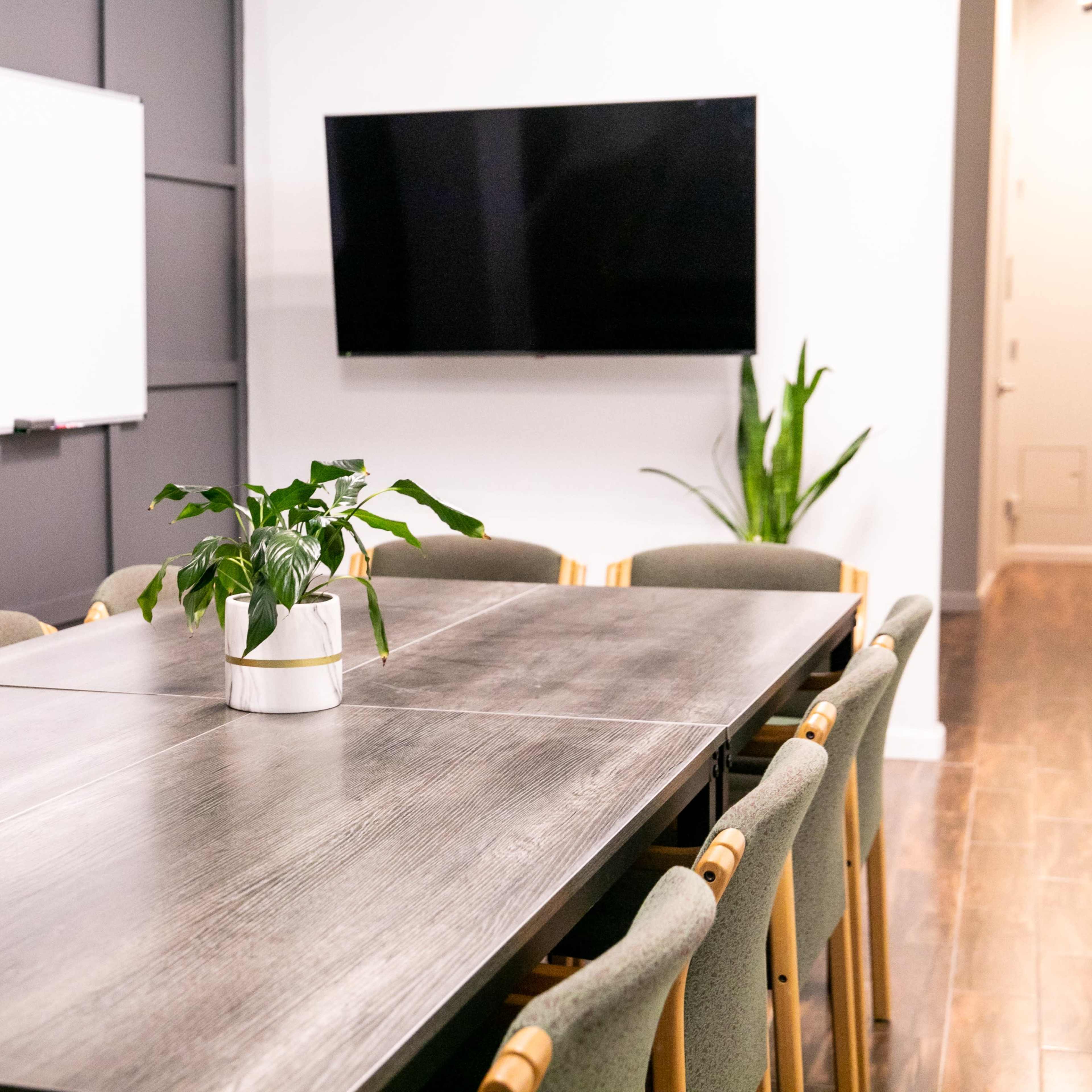 A modern conference room features a large wooden table surrounded by chairs, with a plant centerpiece, a whiteboard, and a television on the wall.