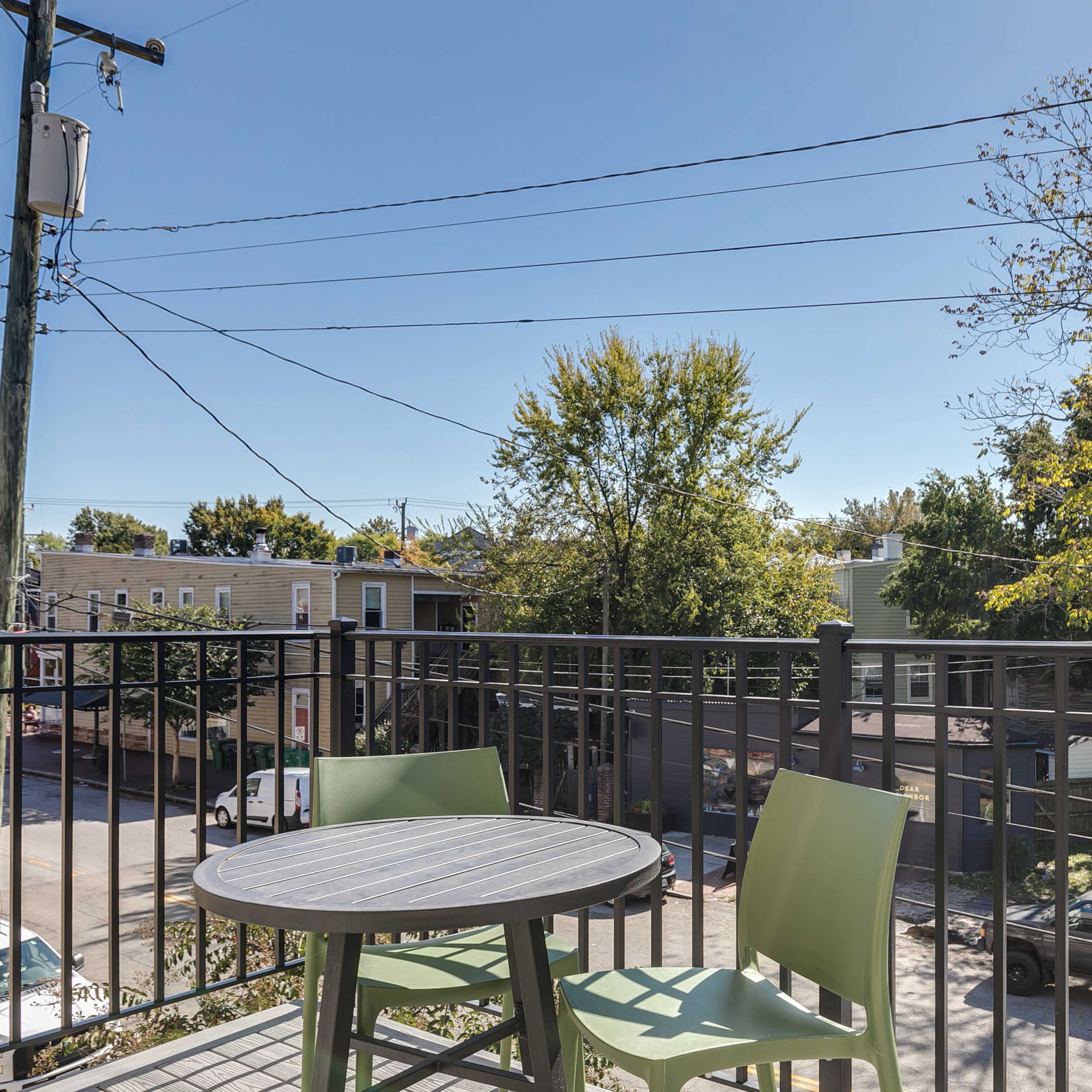 A balcony view features a small round table and two chairs overlooking a tree-lined street with utility poles and parked cars.