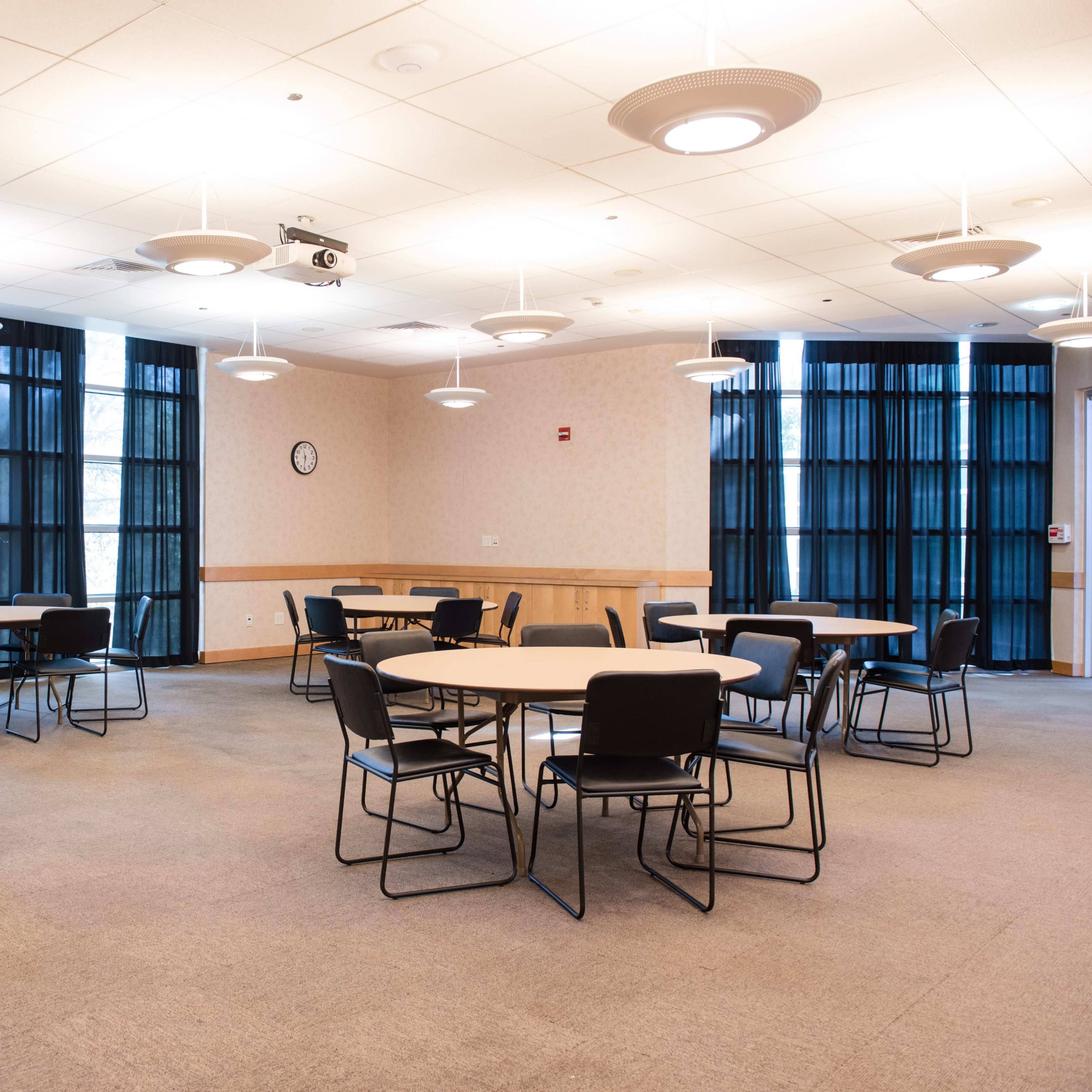 A conference room featuring several round tables and chairs with large windows covered by dark curtains.