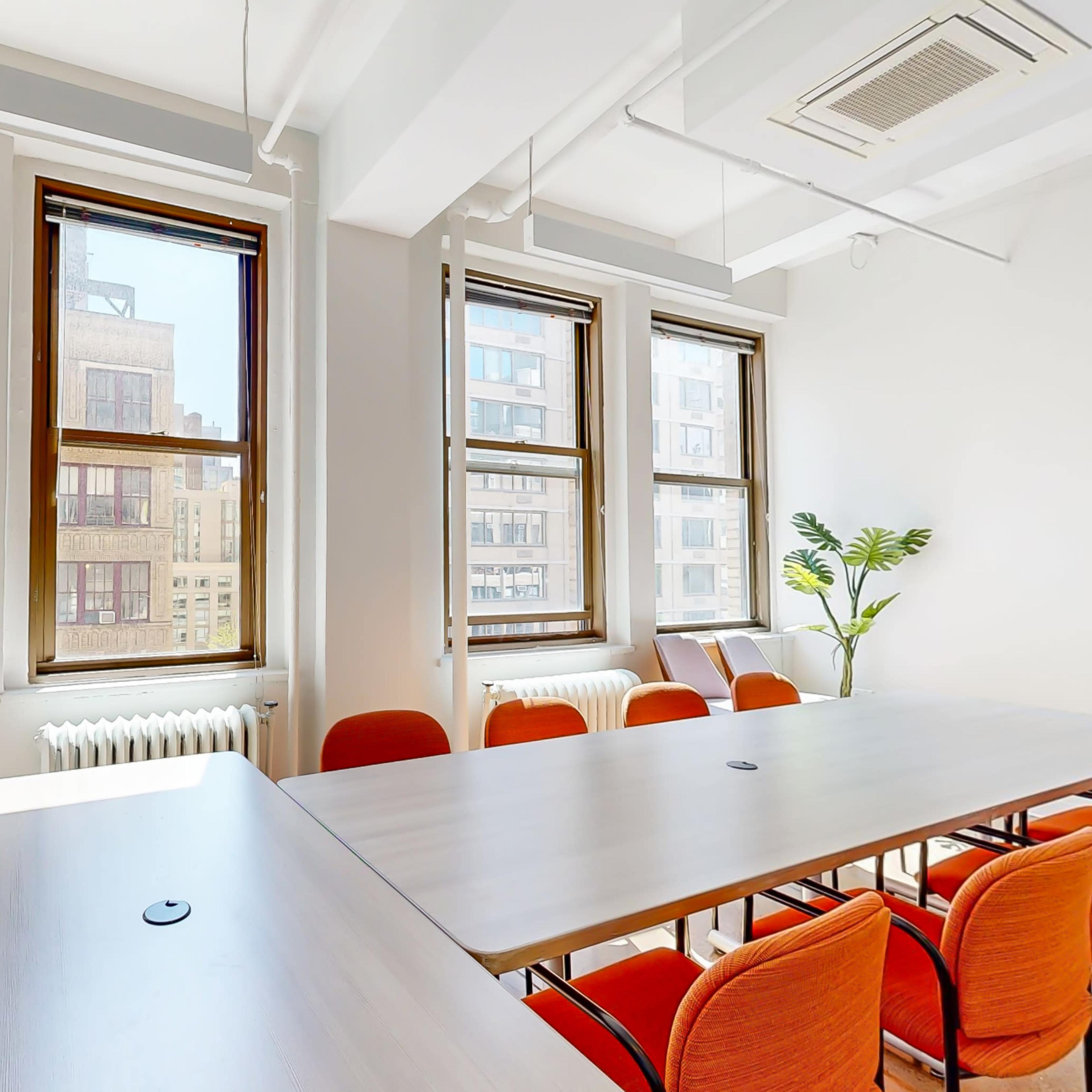A bright meeting room features large windows, a long table, and orange chairs arranged around it.