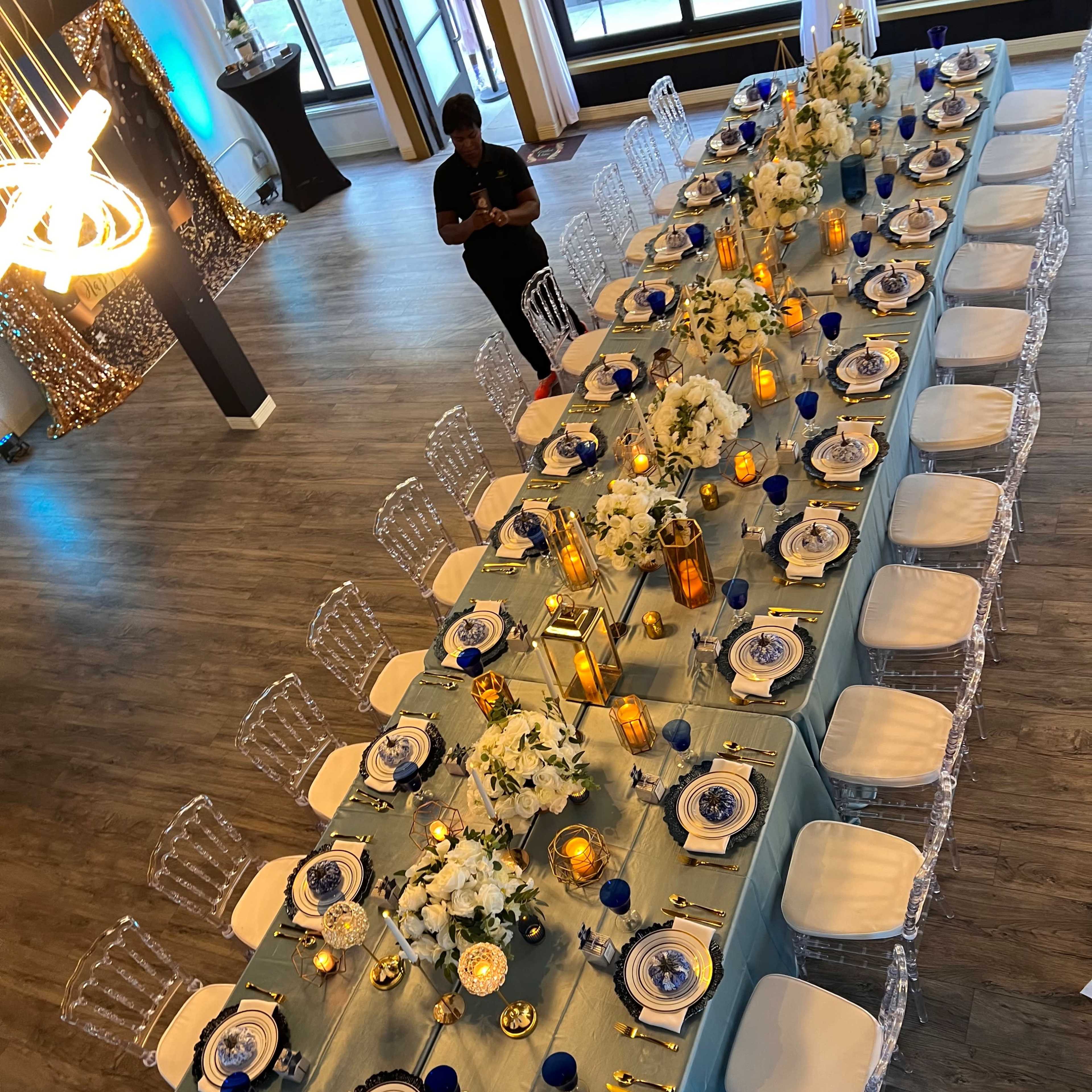 A long banquet table is set with white flowers, blue dishes, and lit candles, surrounded by clear chairs in a spacious event venue.
