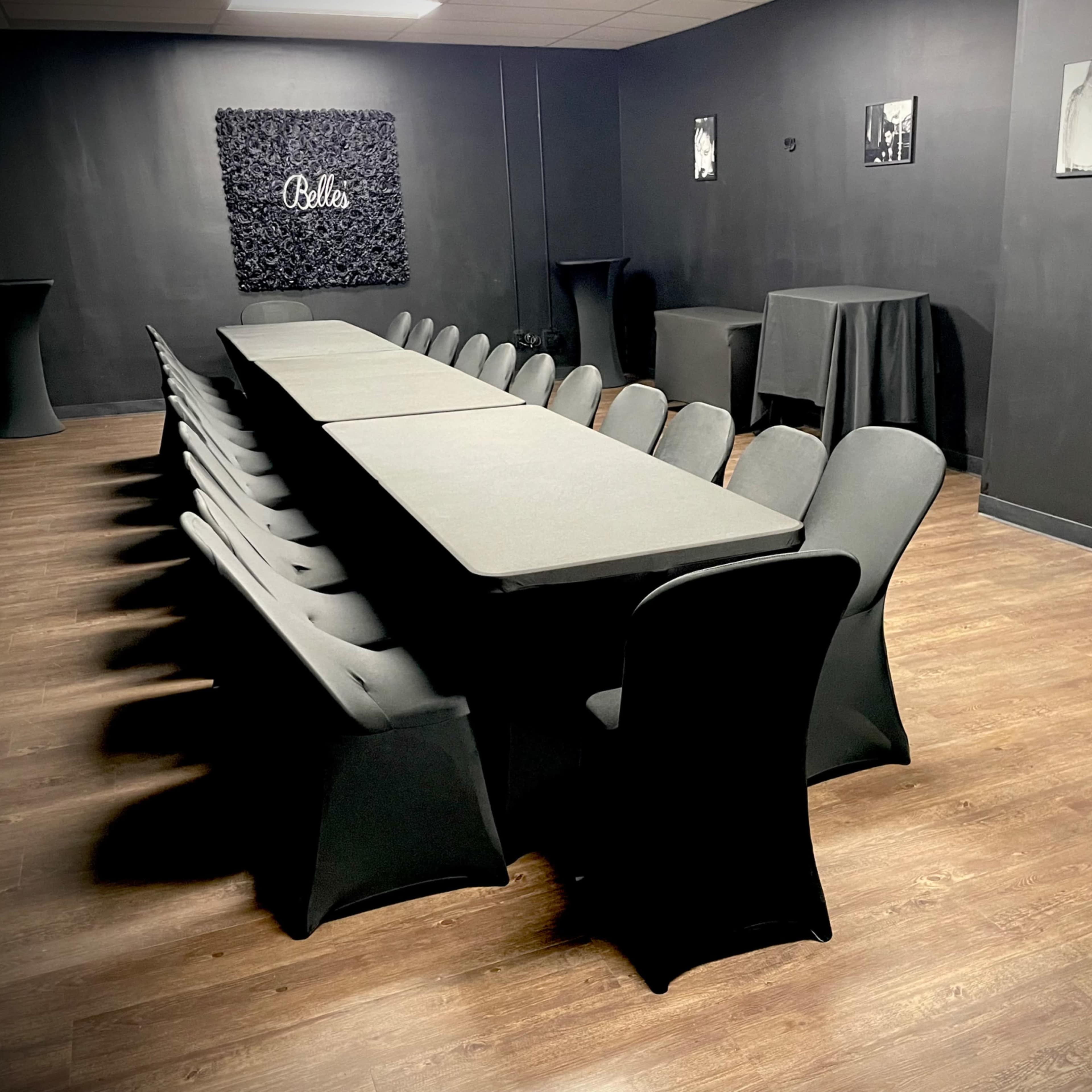 A long rectangular table with black tablecloths is arranged in a dimly lit room, surrounded by chairs, with black walls and a decorative wall featuring the word "Bella."