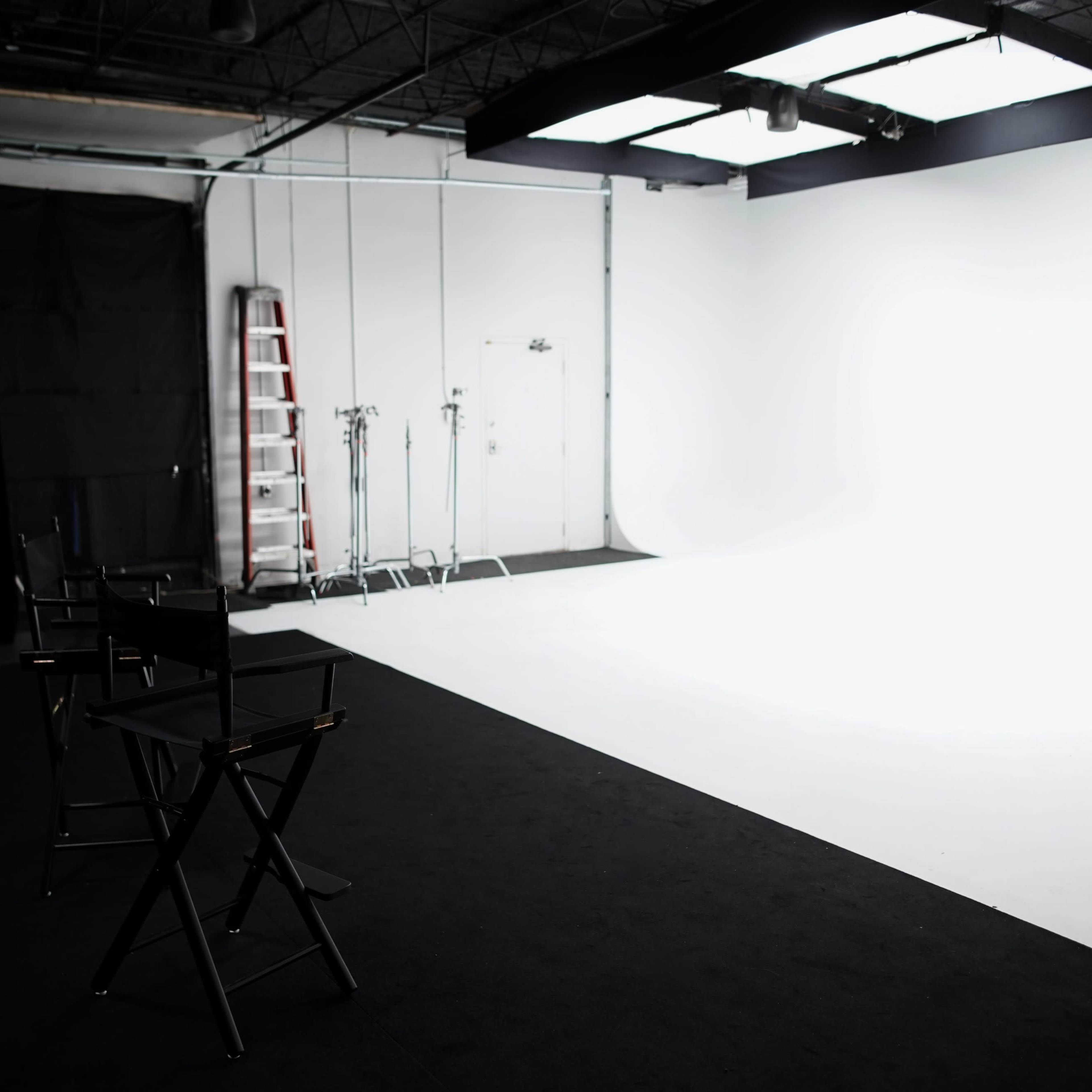 A photography studio features empty black director's chairs facing a blank white backdrop, with a ladder and lighting equipment in the background.