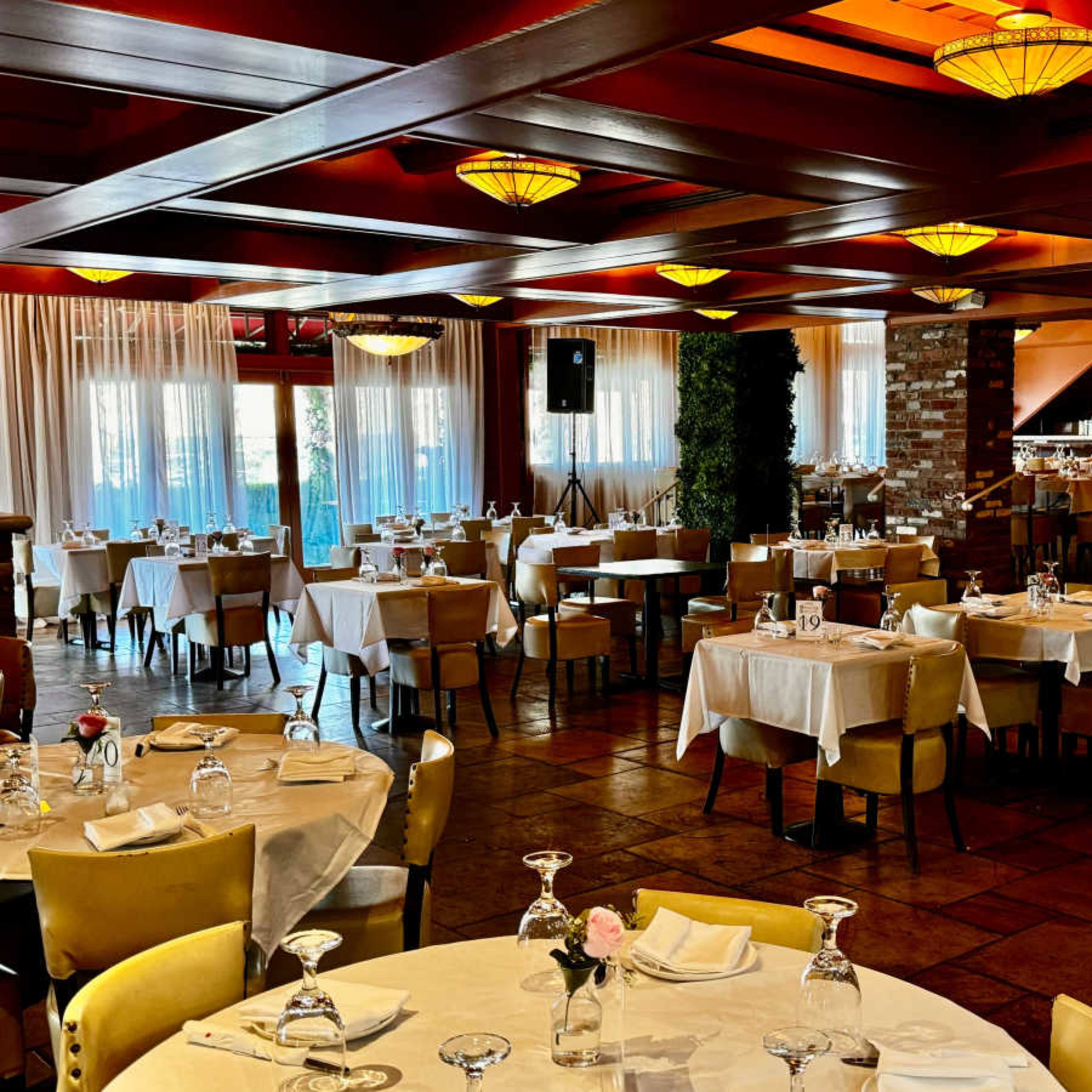 A spacious restaurant interior with multiple dining tables set with glassware and white tablecloths, illuminated by pendant lights.