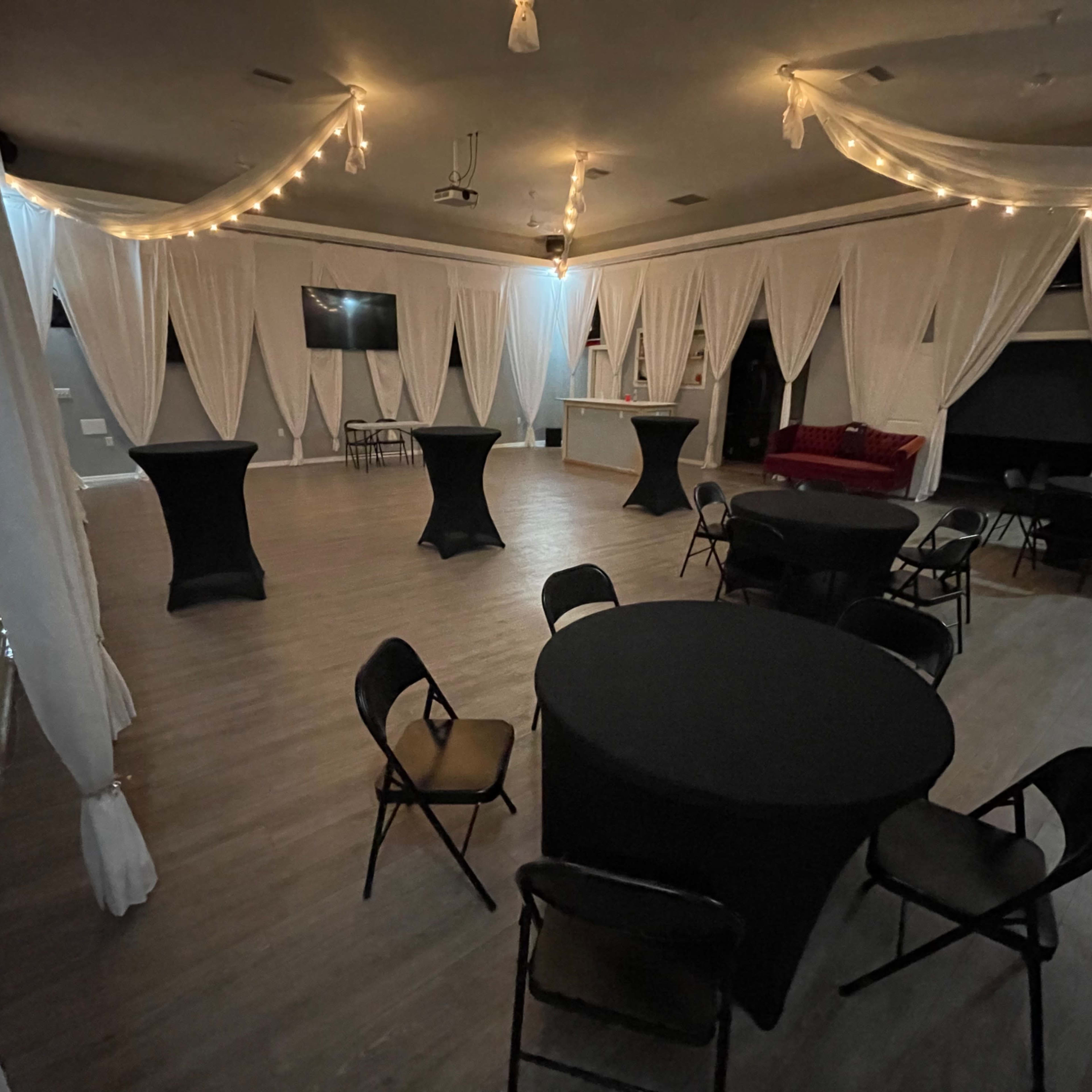 An event space with several round tables covered in black tablecloths, draped white curtains, and lights hanging from the ceiling.