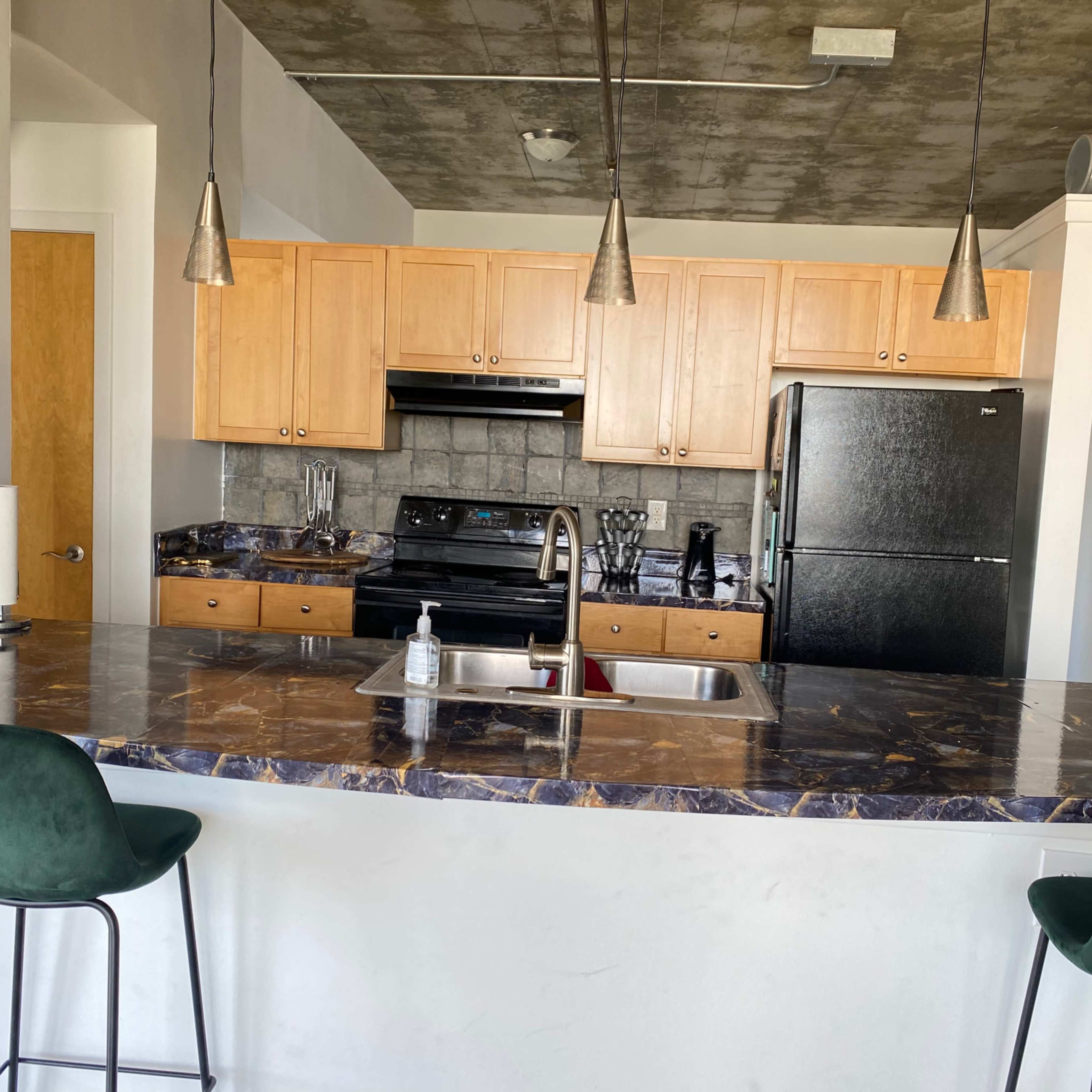 A modern kitchen with wooden cabinets, a black refrigerator, and a marble countertop with two green bar stools.