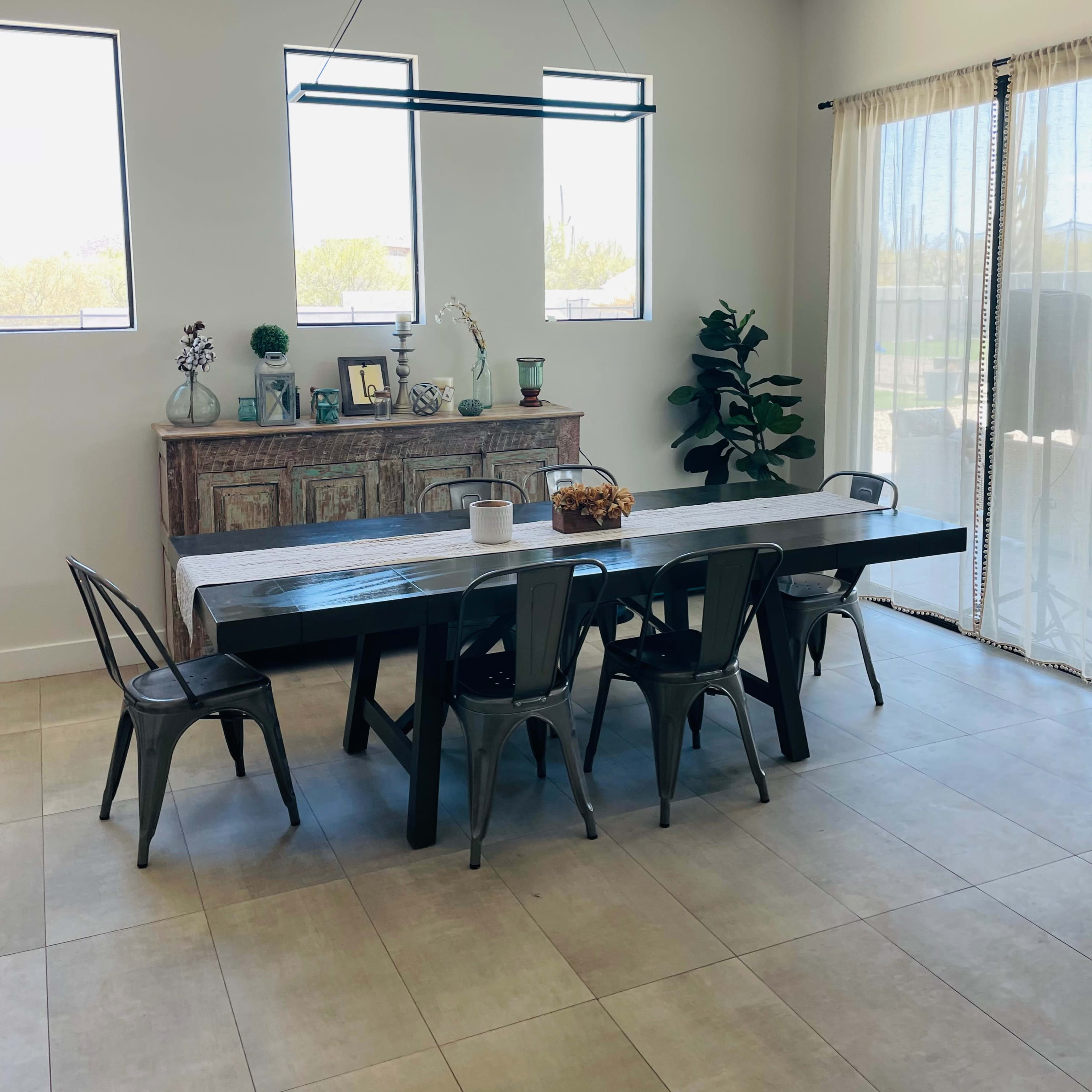 A spacious dining area features a long table surrounded by metal chairs, with a sideboard and plants against the wall, illuminated by natural light from large windows.