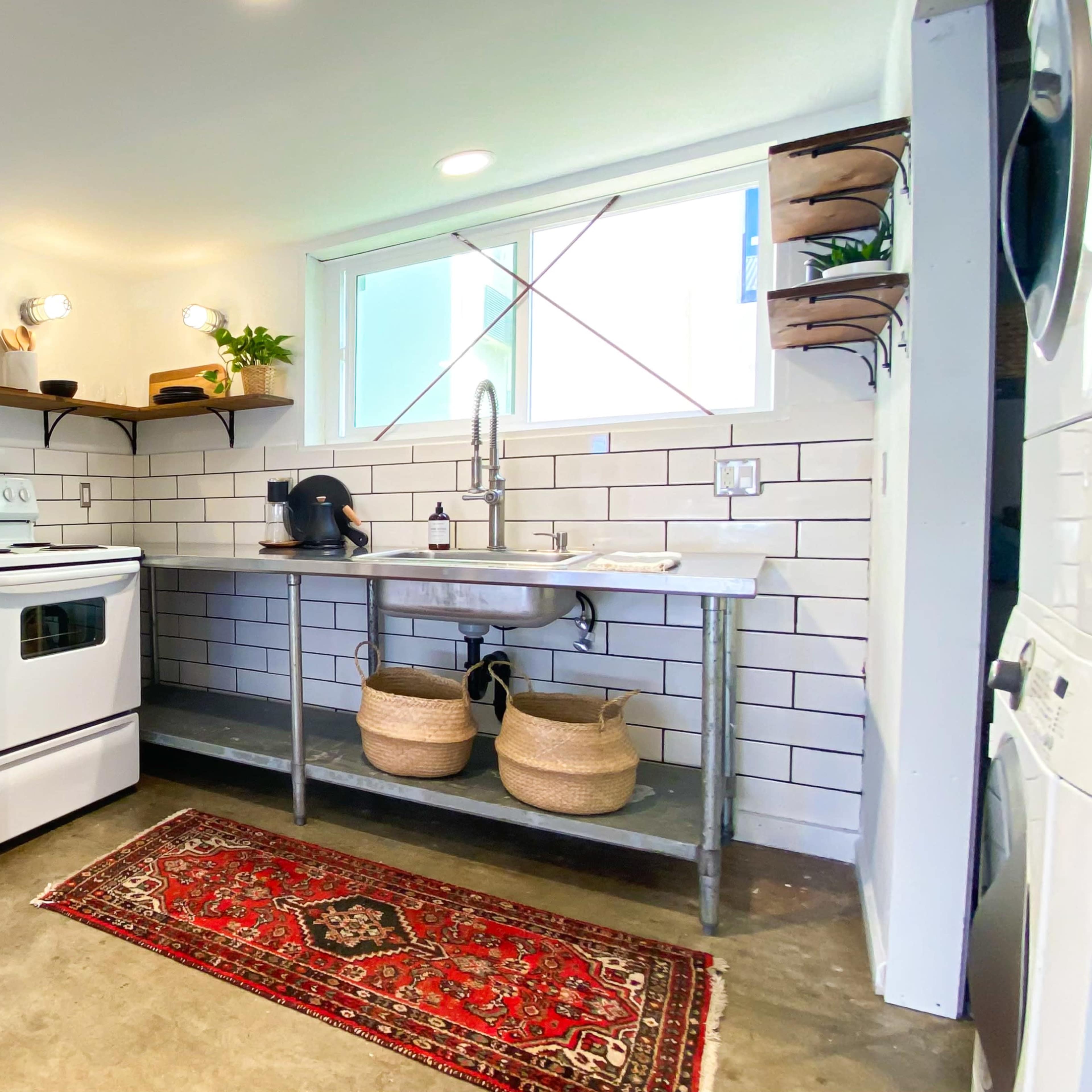 A compact kitchen with a white refrigerator, a stove, a sink on a metal countertop, and decorative elements including a rug and plants.
