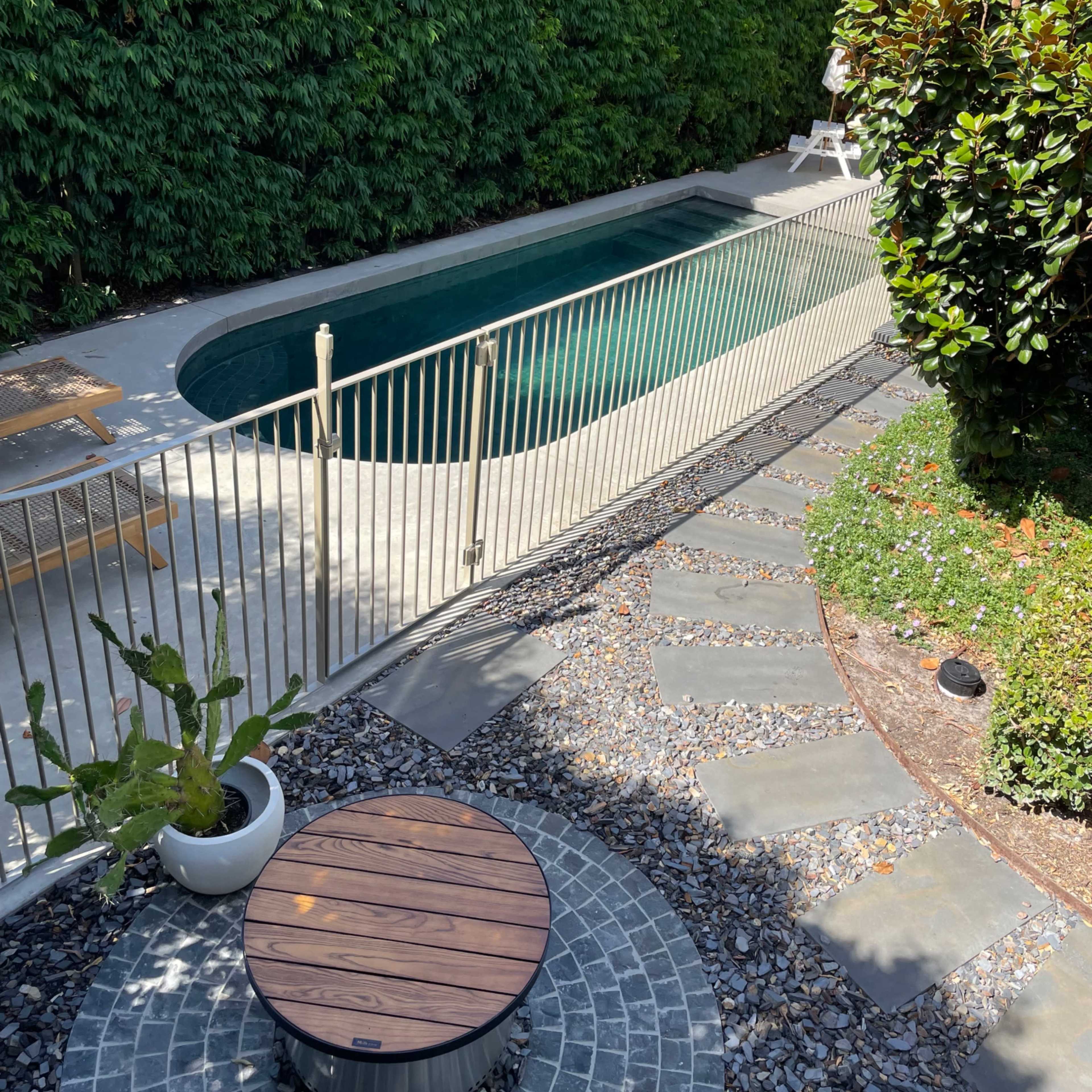A narrow swimming pool enclosed by a metal fence, surrounded by lush greenery and a stone pathway leading to a fire pit area.