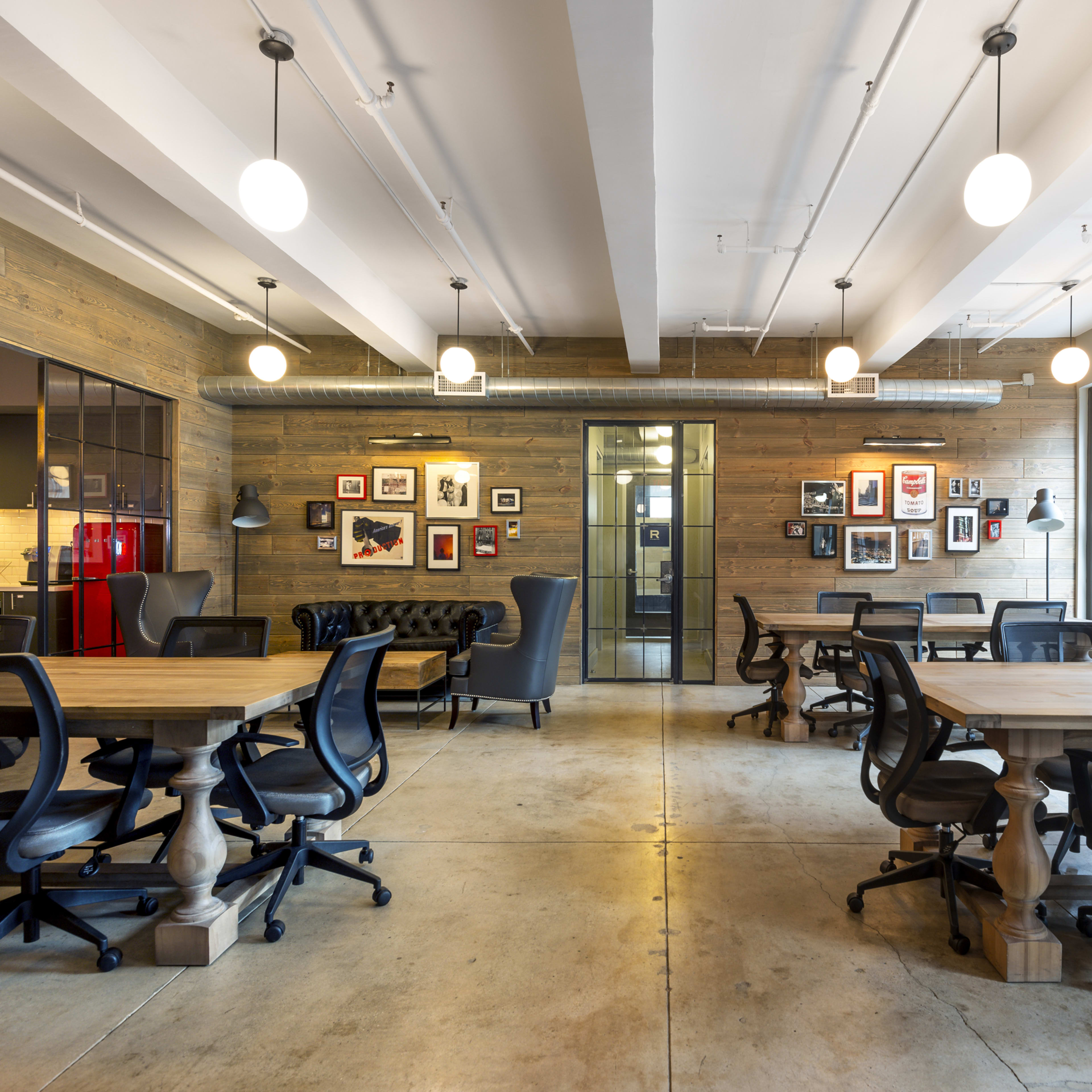 A modern office space featuring multiple wooden tables, ergonomic chairs, and wall decorations with framed photographs.