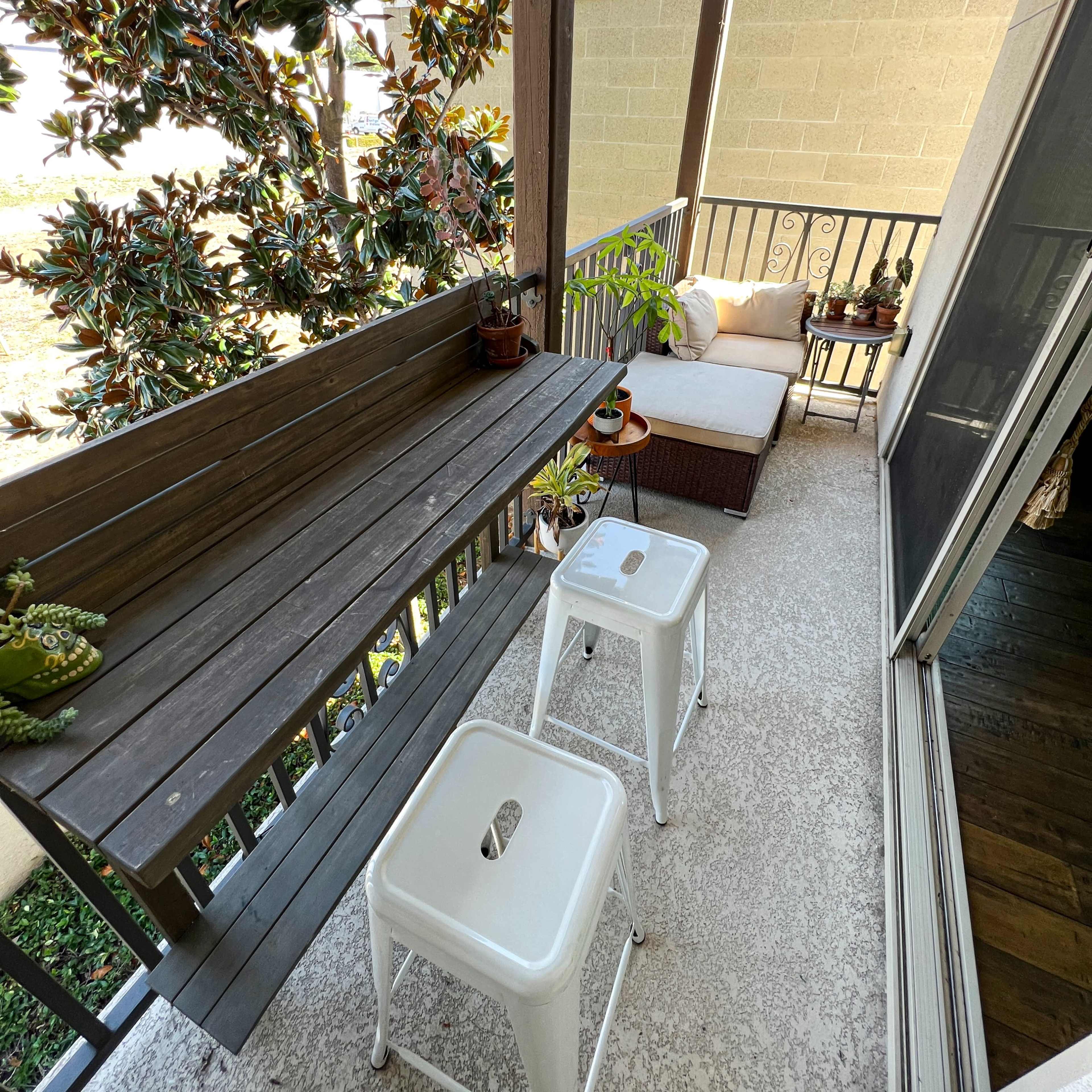 A covered balcony features a wooden bar with two metal stools, plants, and a seating area with a cushion.