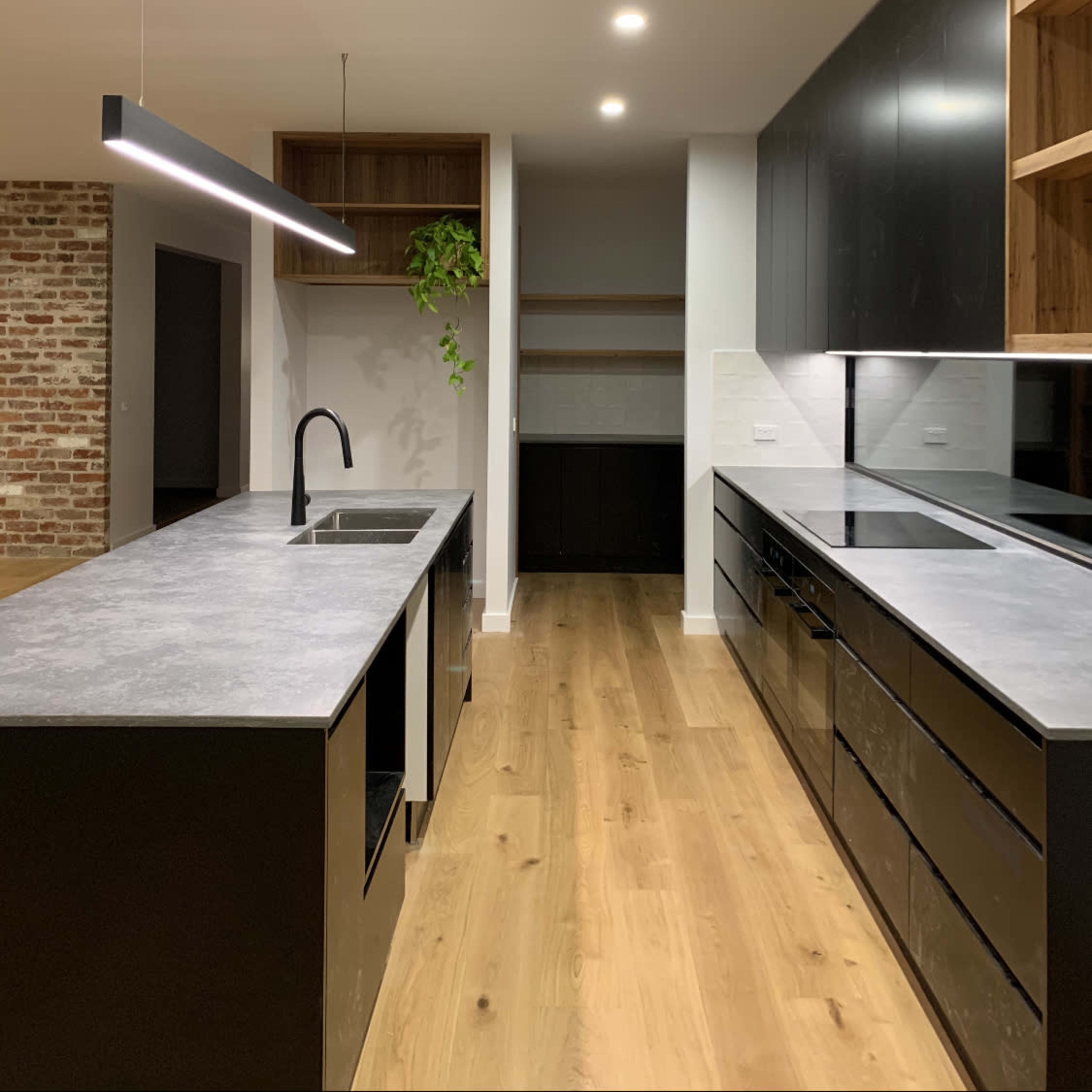 A modern kitchen with a central island featuring a sink, black cabinetry, wooden shelves, and an exposed brick wall.