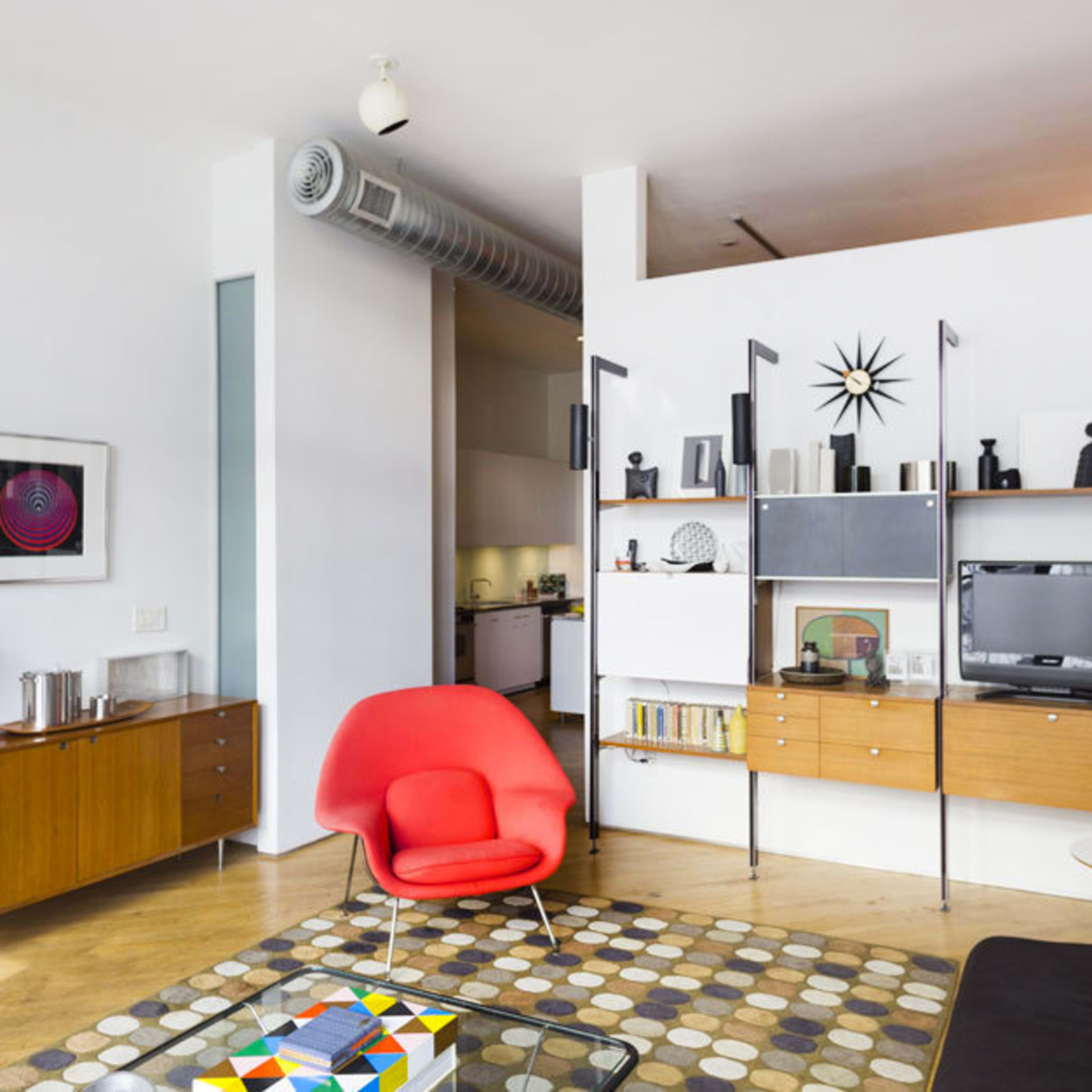 The scene depicts a modern living room featuring a red chair, a wooden sideboard, a television on a shelving unit, and a patterned rug on the wooden floor.