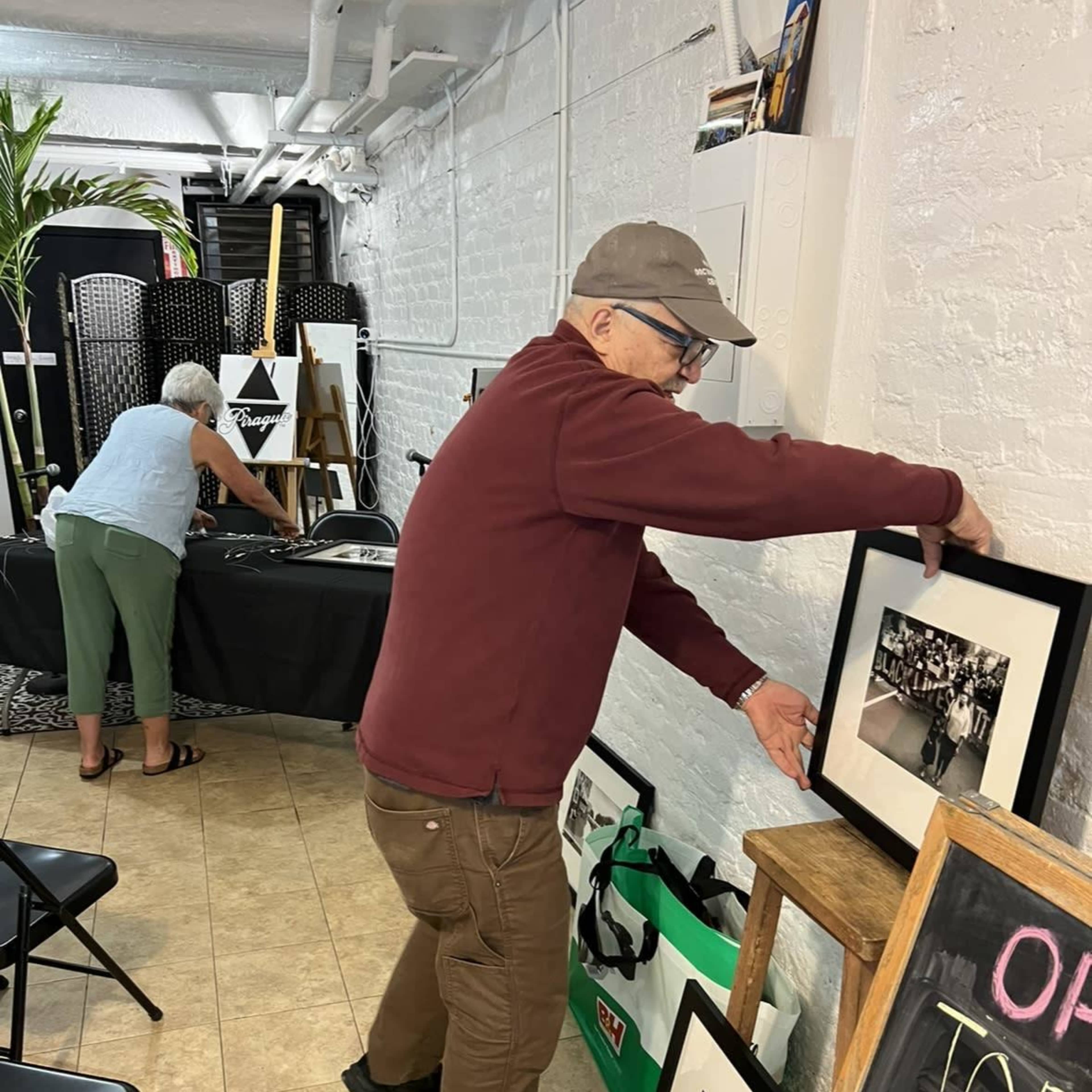 A man adjusts a framed photograph on a display while a woman organizes items on a table in a gallery-like space.