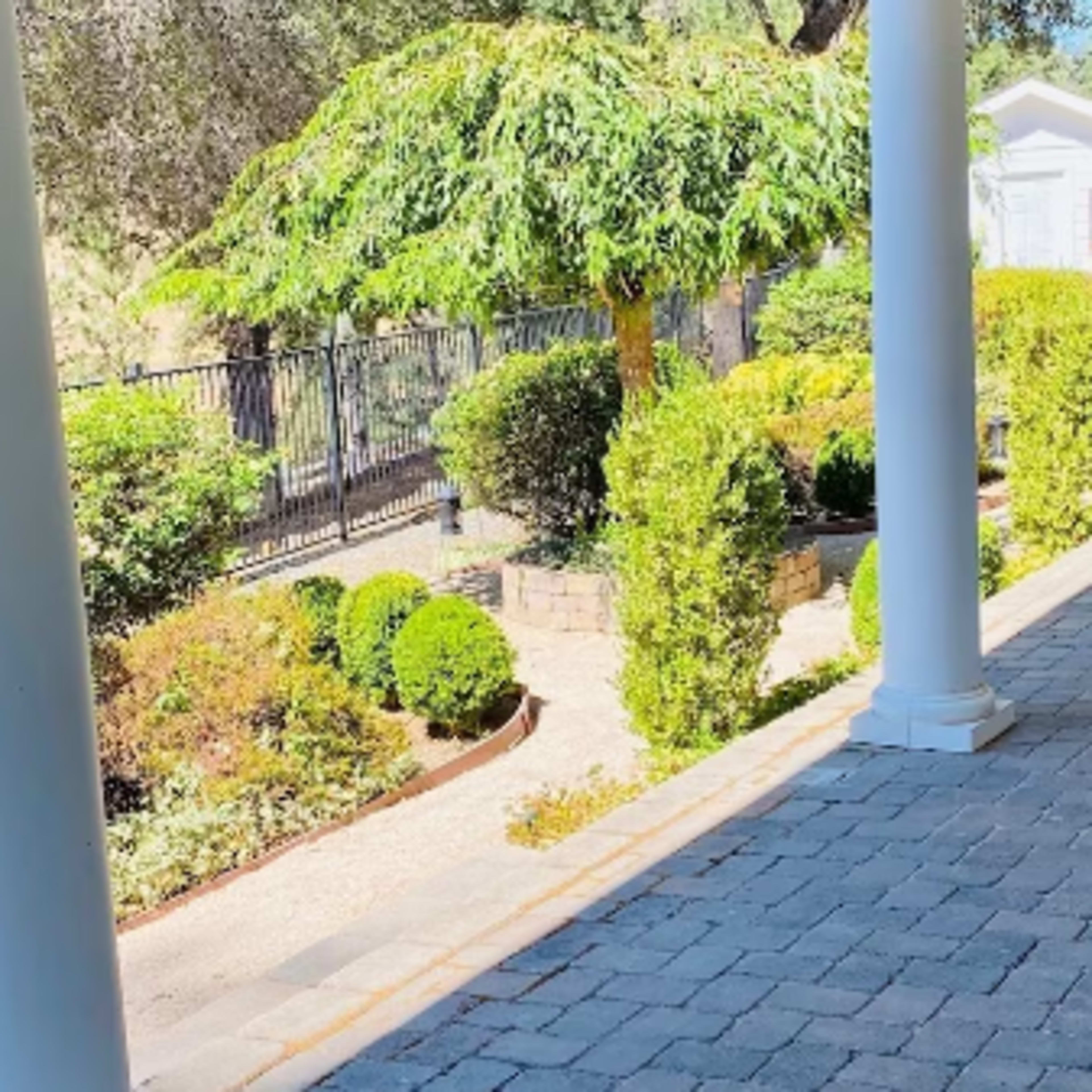 A covered patio features a stone floor, surrounded by neatly trimmed bushes and trees, with a dining set in the background.