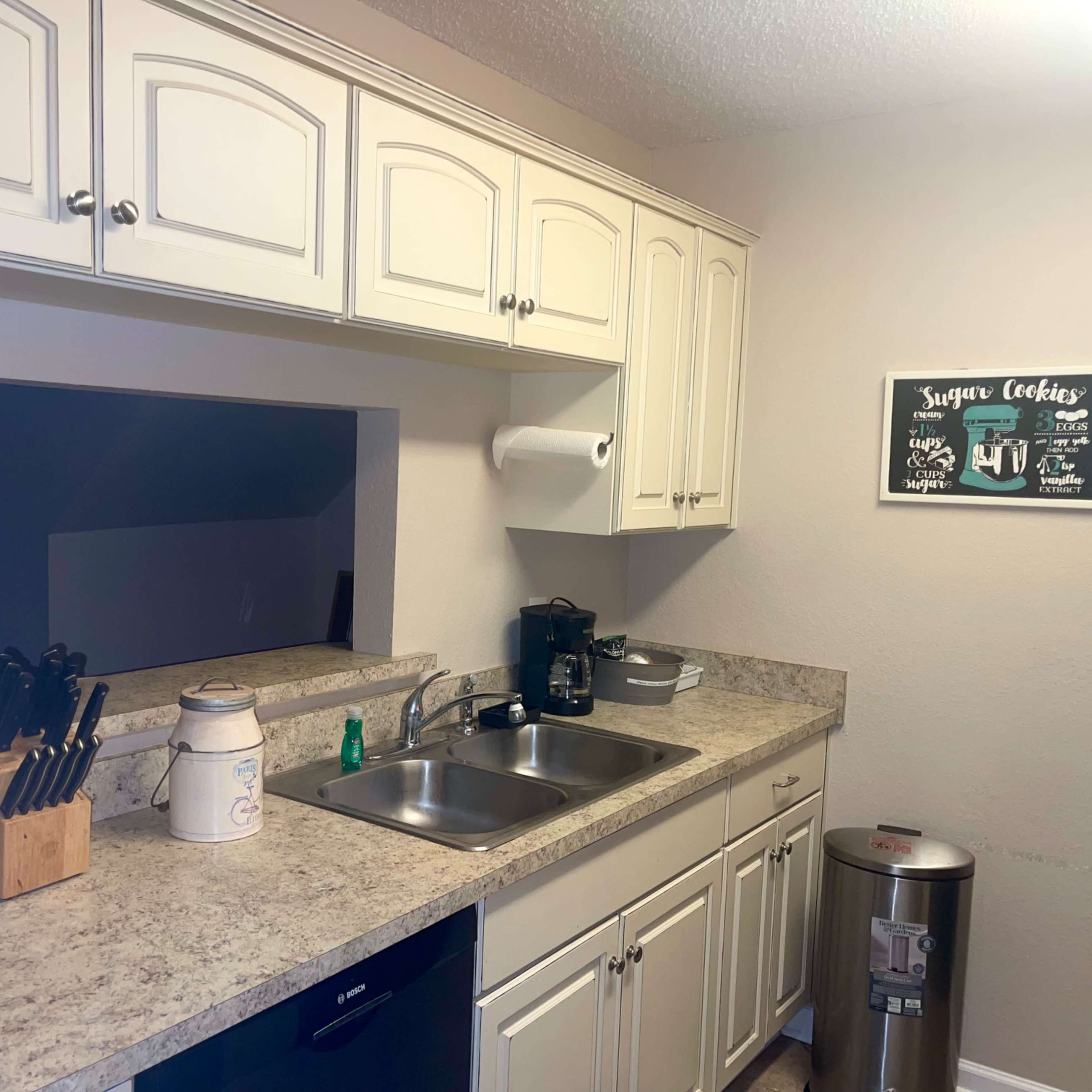 A small kitchen features white cabinetry, a double sink under a window, and various appliances, including a coffee maker and a trash bin.