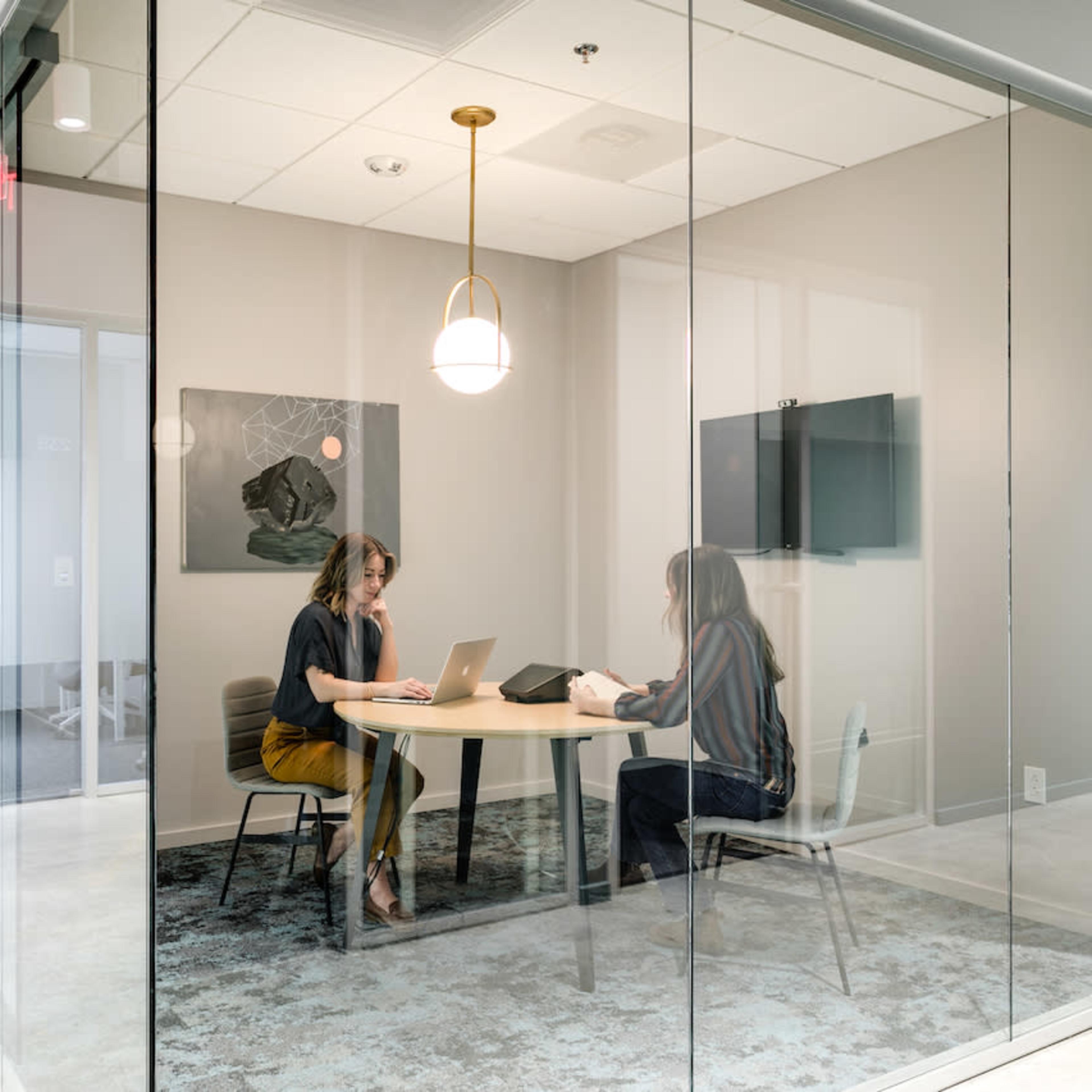 Two women are sitting at a table in a glass-walled meeting room, using laptops and tablets.