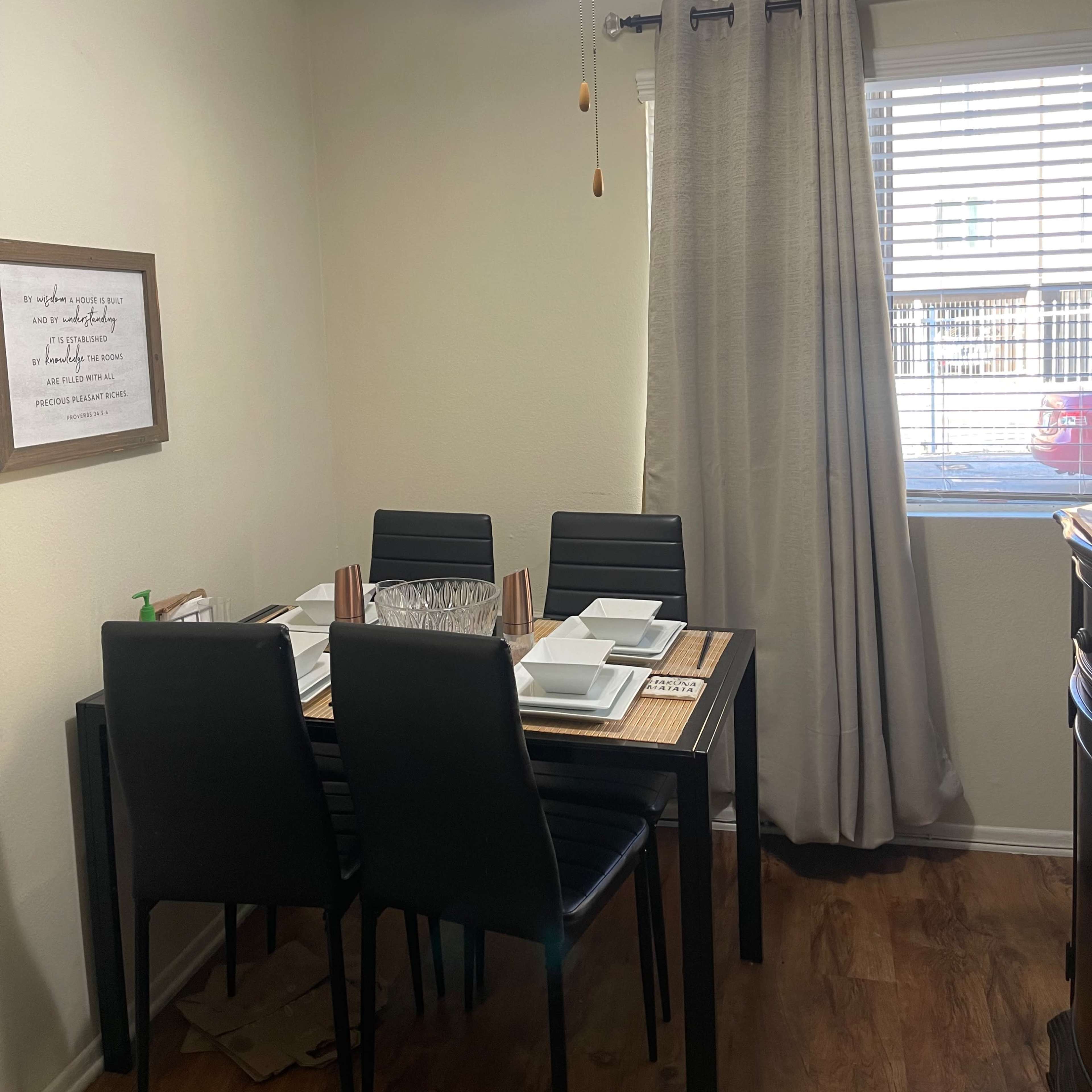 A dining area features a black table with six chairs set with plates, a ceiling fan above, and a window with curtains.