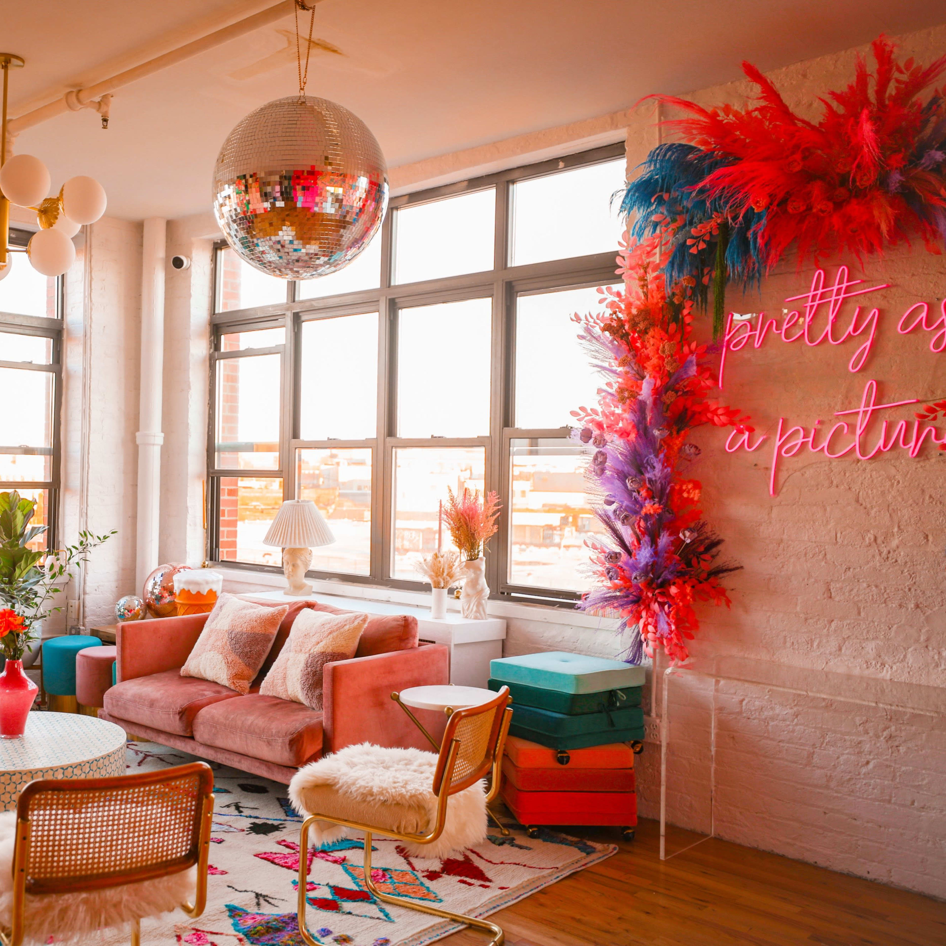 A brightly lit living room with large windows, colorful furniture, a disco ball, and decorative plants.